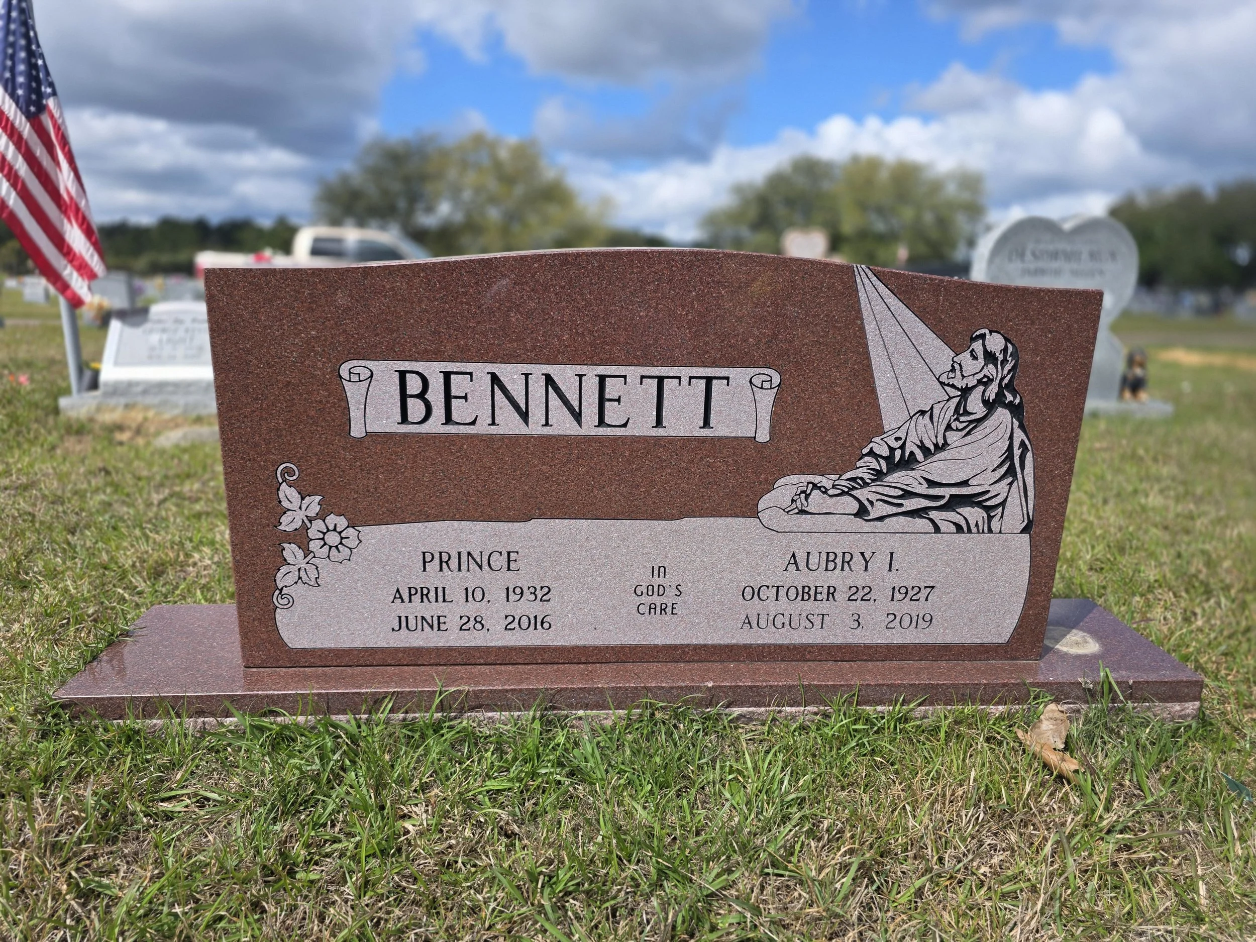 Gravestone with the name Bennett, featuring an illustration of Jesus Christ and a flower branch, with inscriptions for Prince and Aubry I, including their birth and death dates, in a cemetery with other gravestones, grassy ground, and a partly cloudy