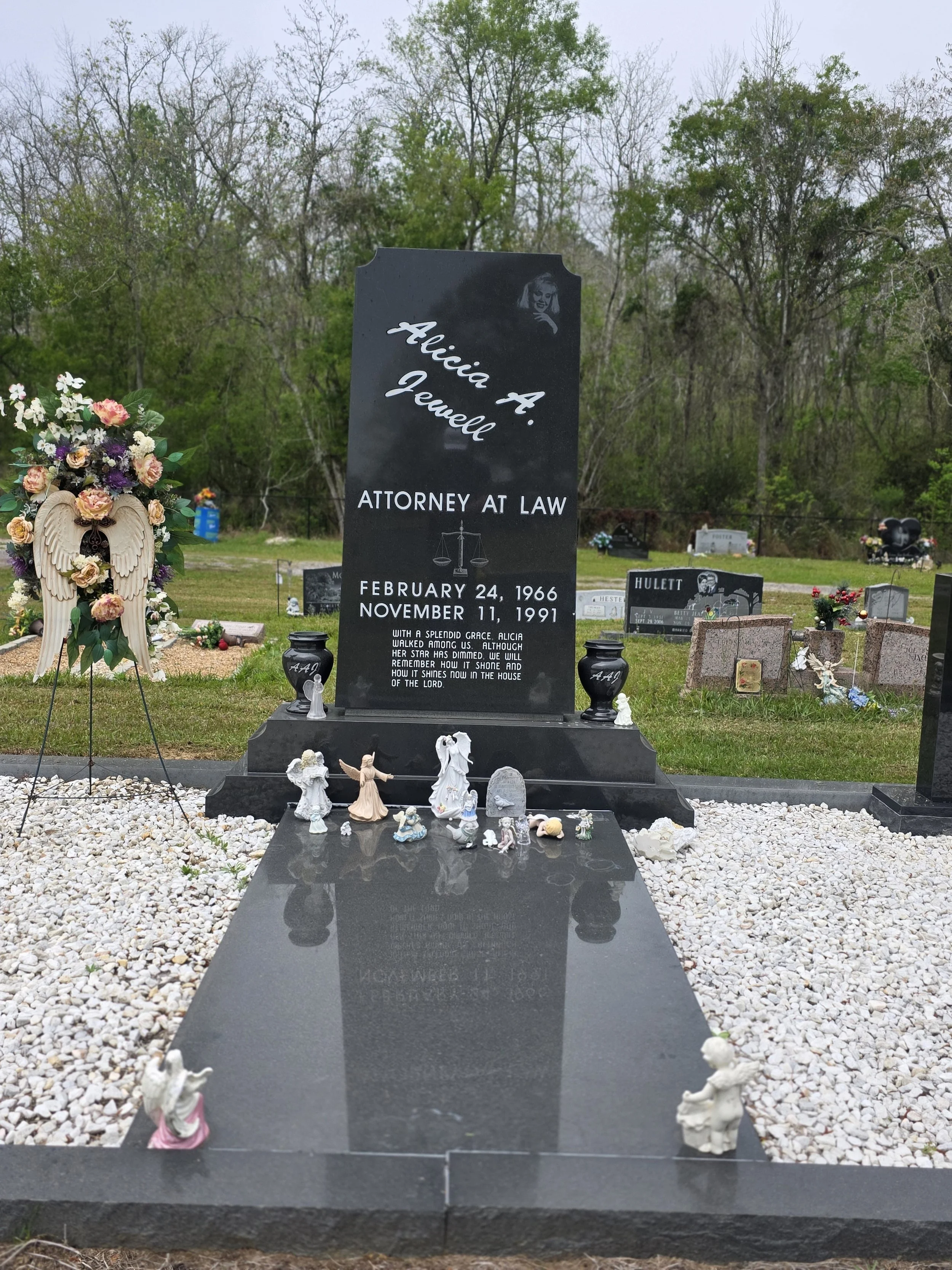 Gravestone with the name Alicia A. Jewell, who was an attorney at law, with dates February 24, 1966, and November 11, 1991. The grave features small angel figurines, flowers, and a cross, with other graves and trees in the background.