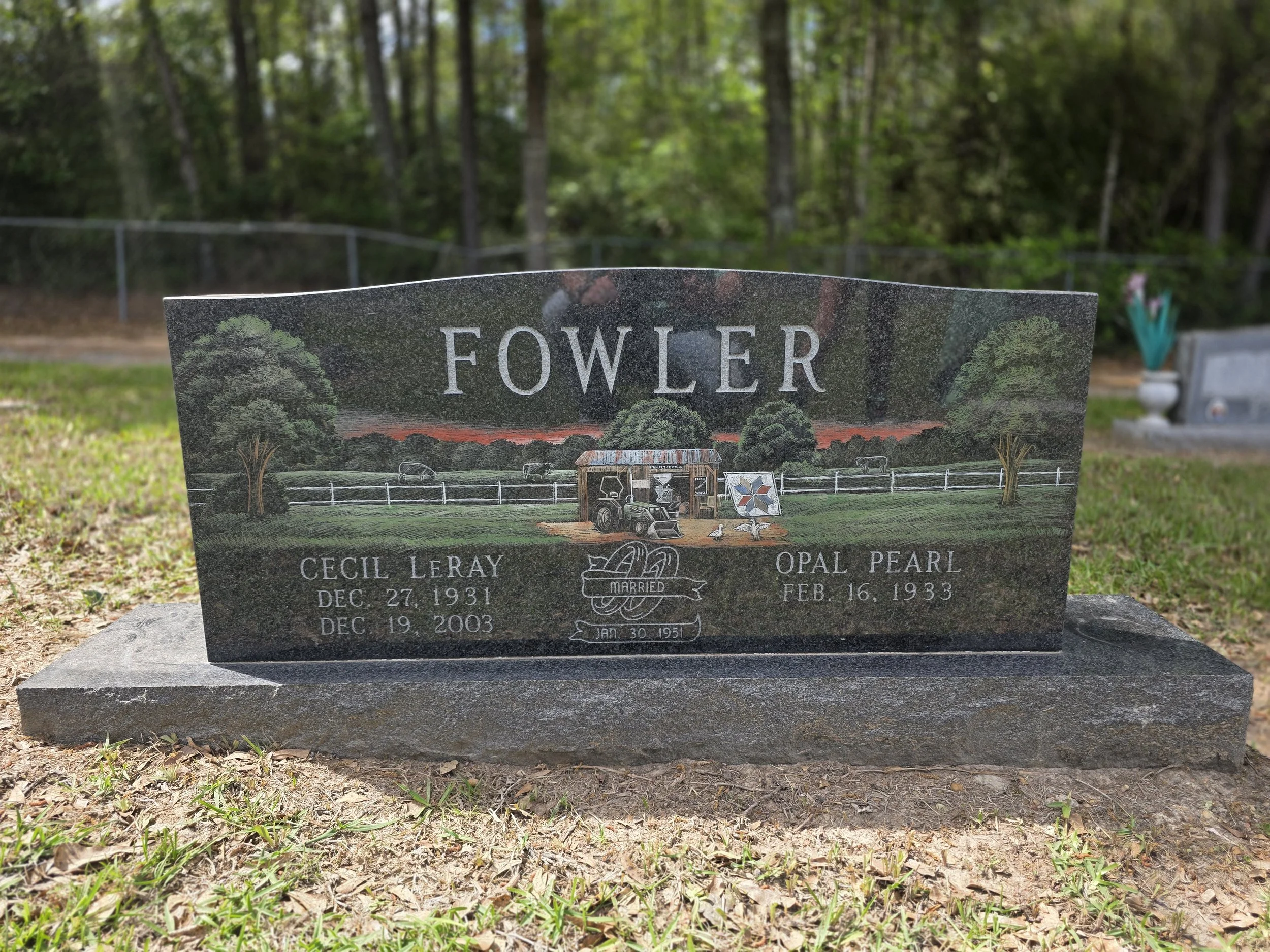 Gray granite headstone with carved trees, fencing, and small farm objects including a horse trailer and a windmill, located in a grassy cemetery.