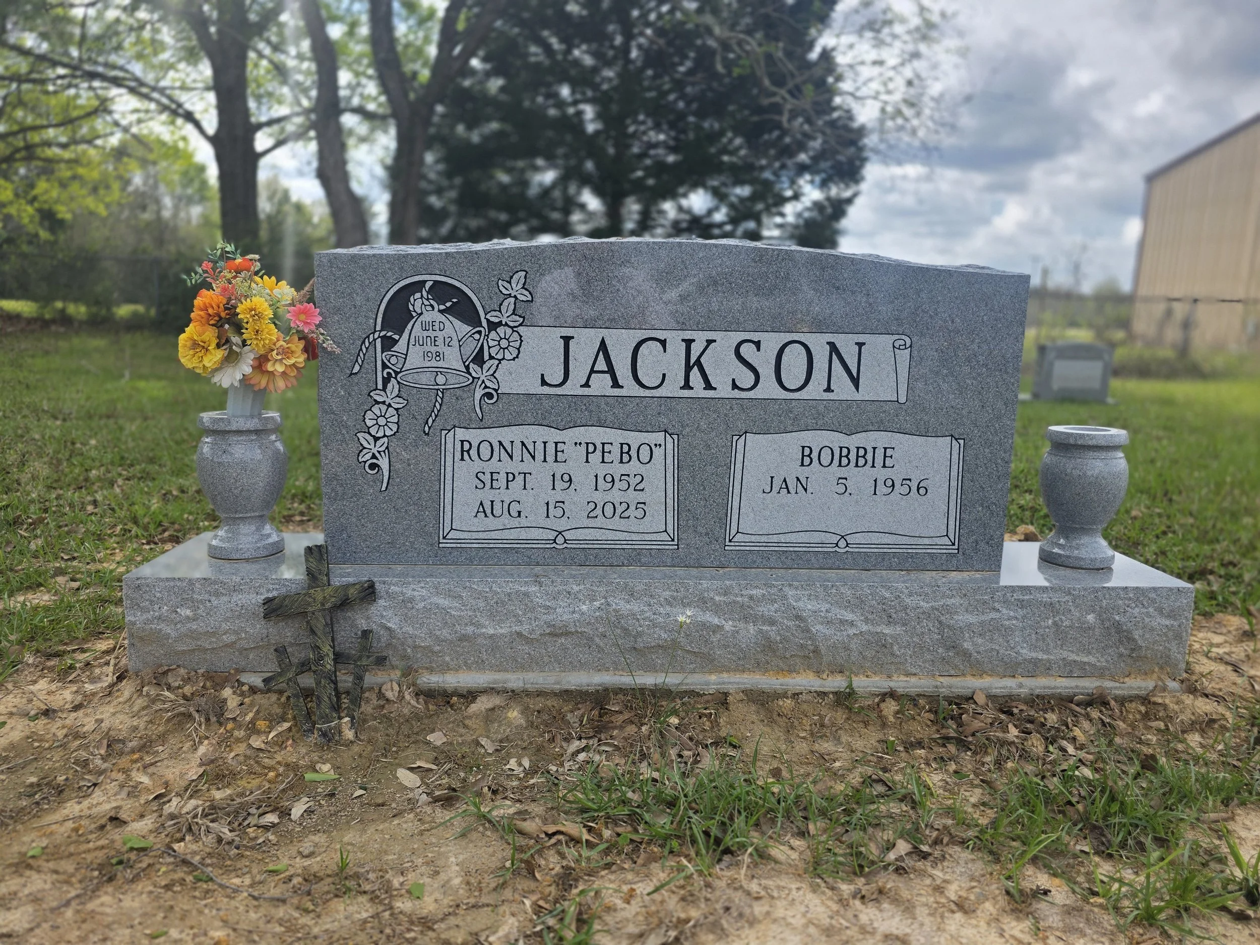 A gray granite tombstone with the name 'Jackson'. It has a floral engraving with a bell that reads 'Wed June 12 1981'. The tombstone includes two names: Ronnie 'Pebo' Jackson (September 19, 1952 - August 15, 2025) and Bobbie Jackson (January 5, 1956)