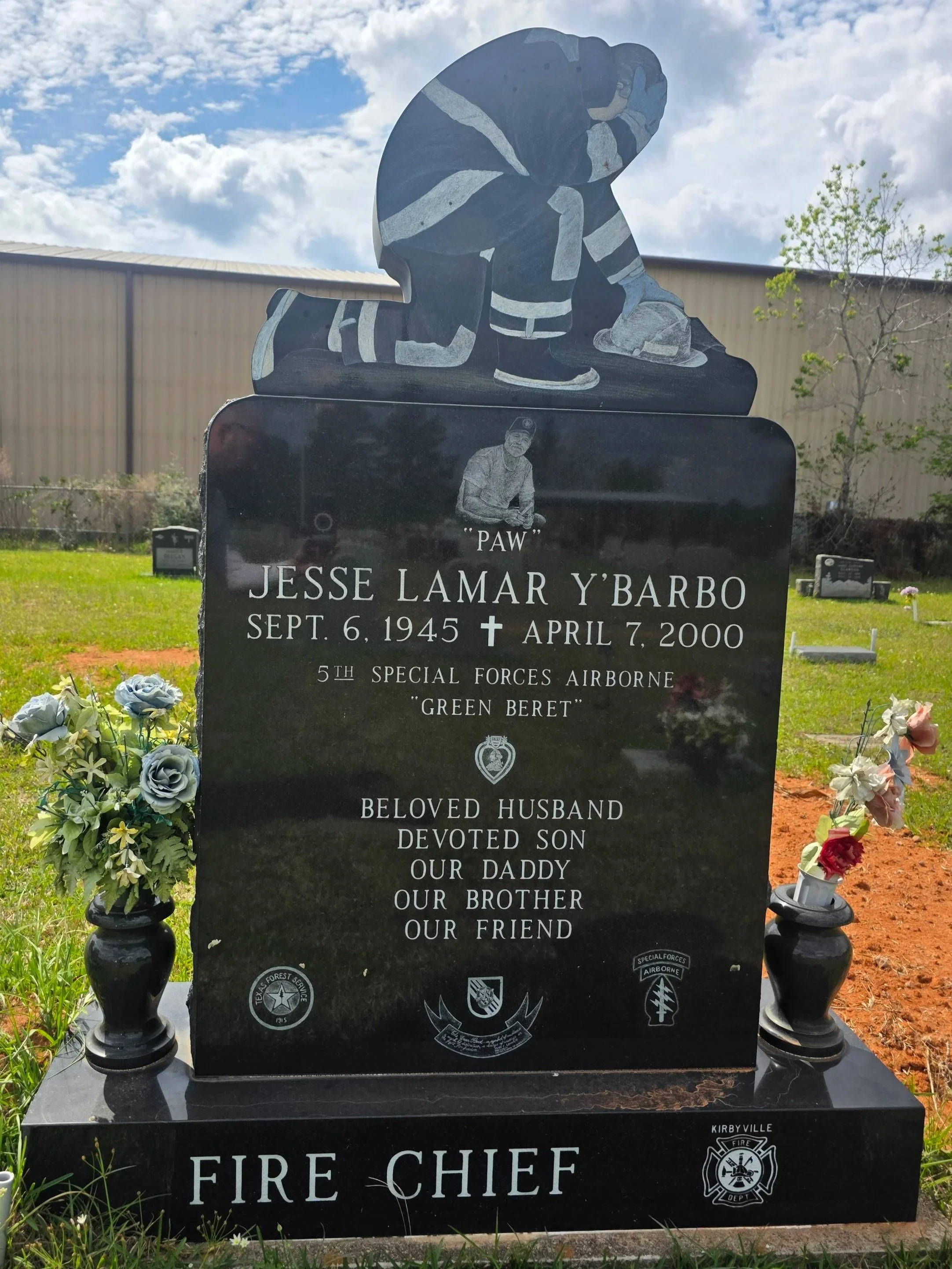 Gravestone with a painted firefighter sculpture on top. The headstone is black and features inscriptions