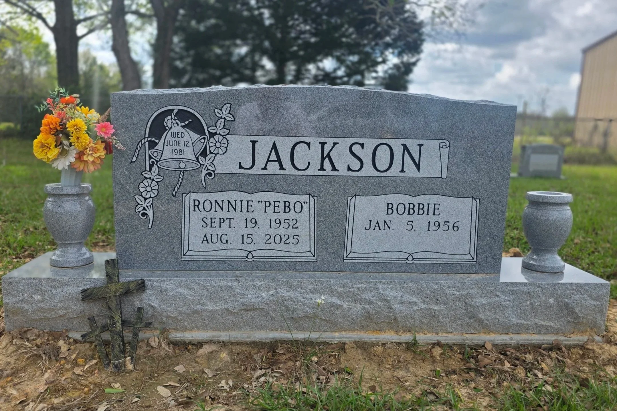 A gray granite headstone There are two vases with colorful flowers on either side of the headstone, and a small cross at the front of the grave.