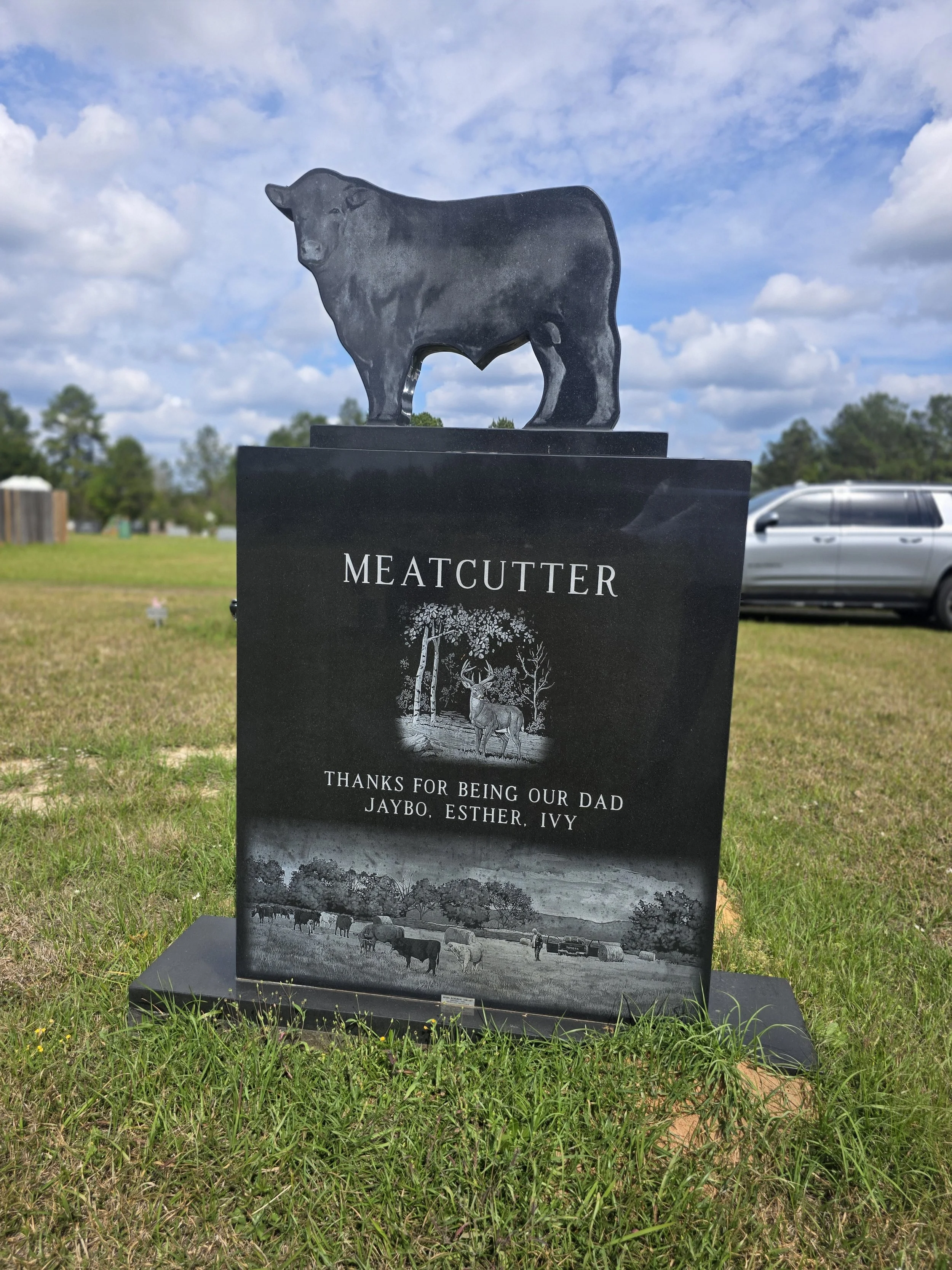 A black tombstone with a bear sculpture on top, inscribed with 'MEATCUTTER' and a message thanking for being a dad to Jaybo, Esther, and Ivy. The tombstone features a rural landscape with cows and trees in the background.