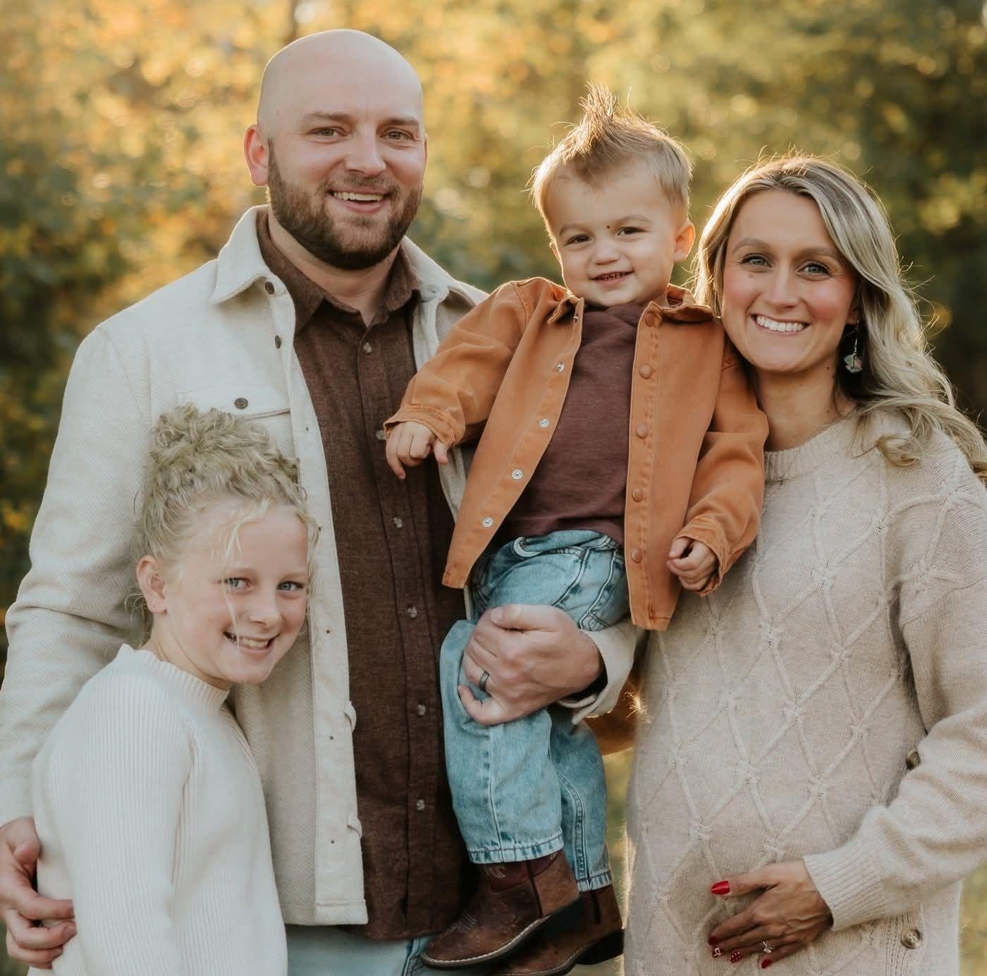 A happy family of five outside in autumn, with a bald man holding a young boy, a woman with long blonde hair, and a girl with curly blonde hair all smiling.
