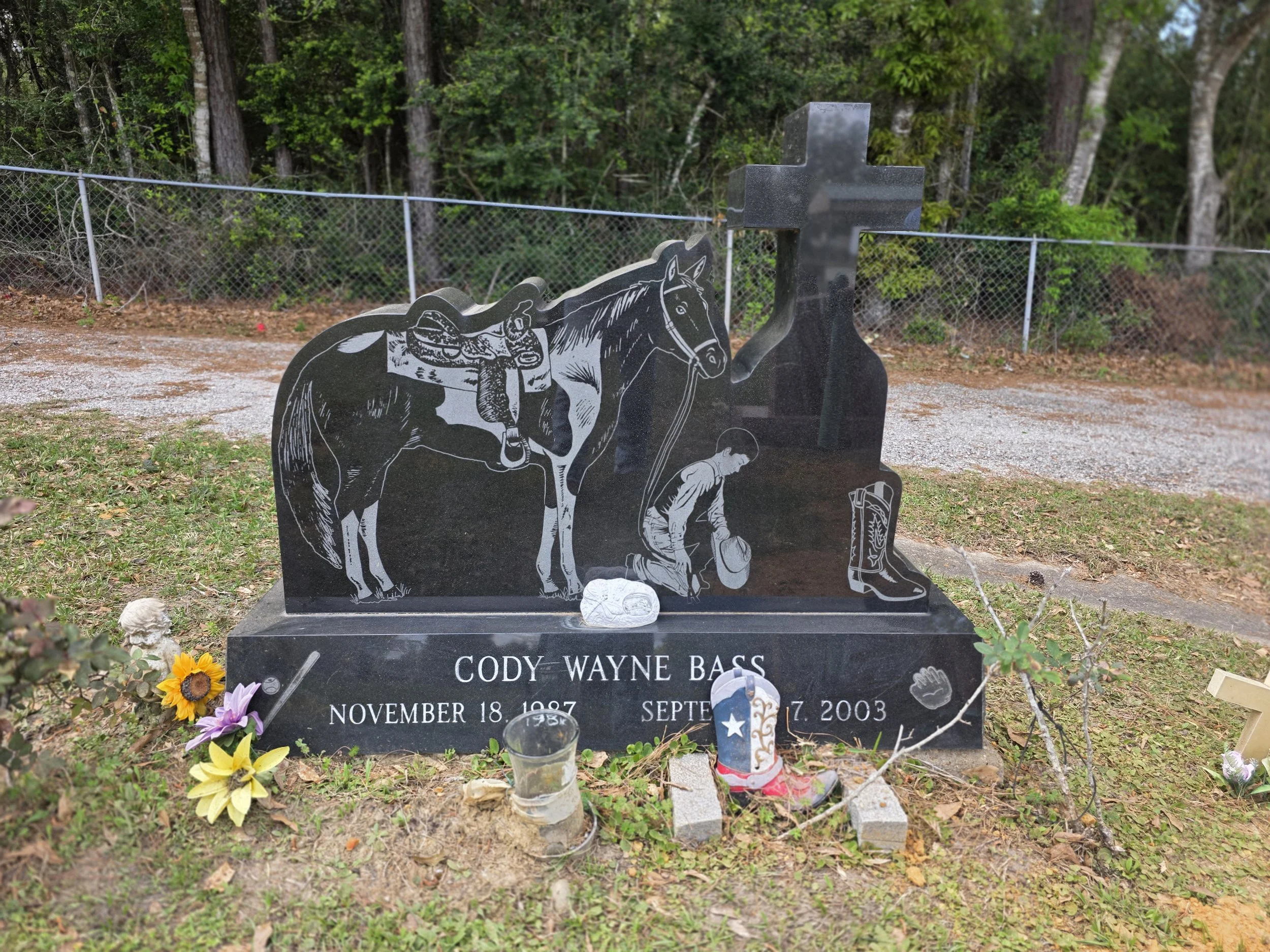 Gravestone with etched images of a horse, a cowboy, and a lone boot, with flowers and candles in front, in a grassy area surrounded by trees and a chain-link fence.