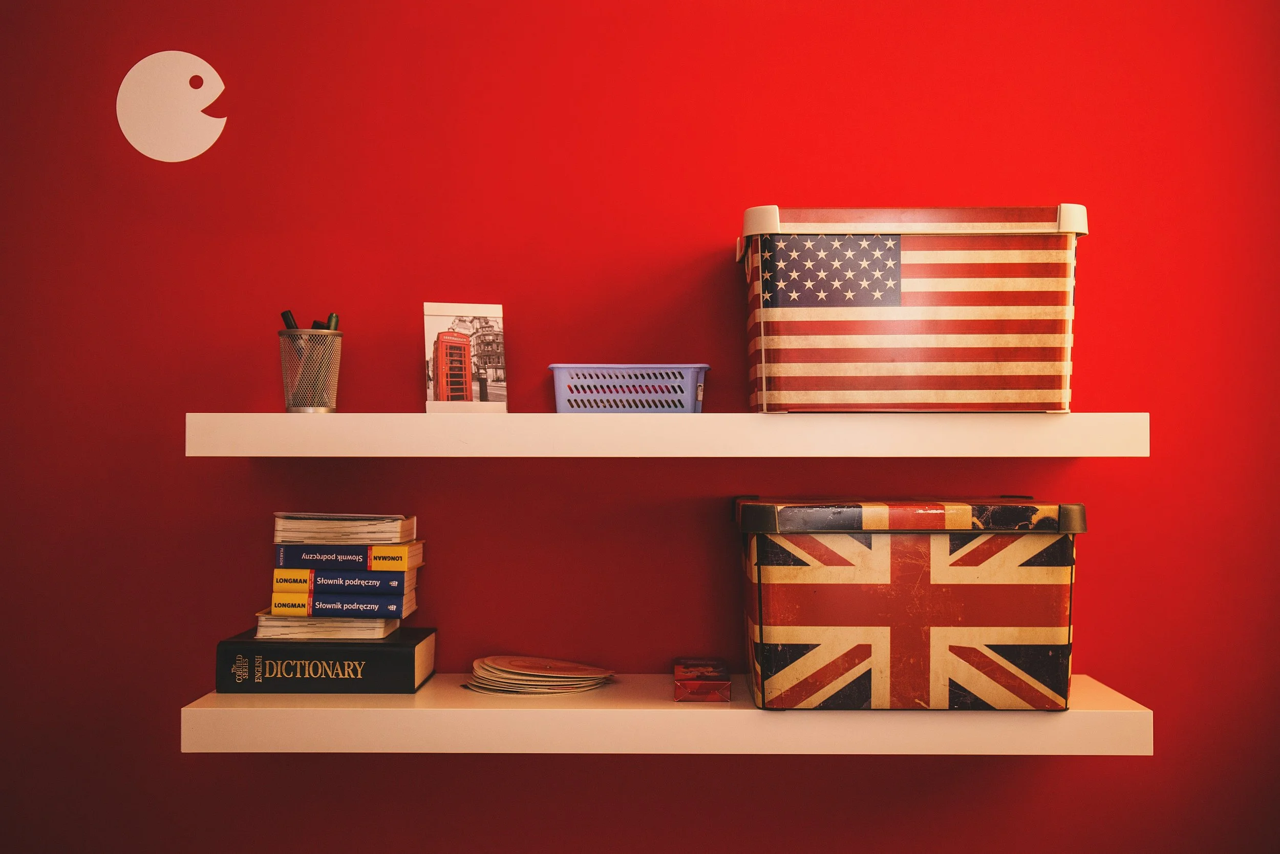 Two white floating shelves with British and American themed storage boxes, books, a framed photo, a stack of books, and small containers against a red wall, with a Pac-Man graphic in the upper left corner.