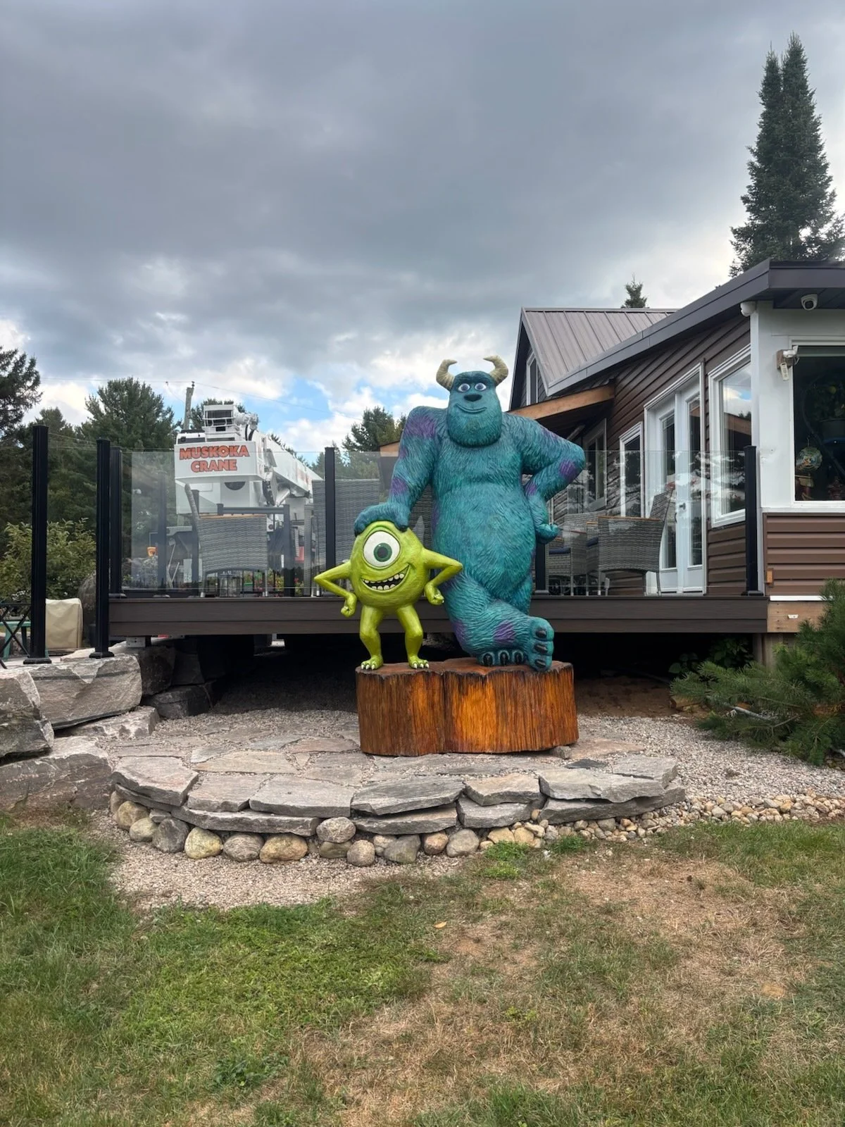 Colorful statue of Mike and Sulley from Monsters Inc. on a wooden base in front of a house with a deck, trees, and cloudy sky.