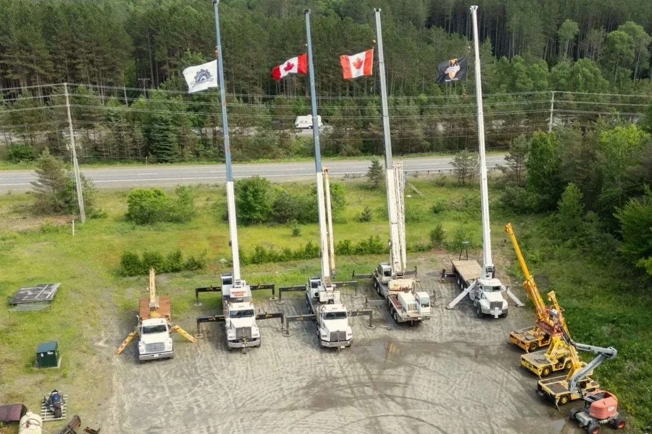 Five flagpoles with flags, including Canada, and construction equipment parked on a gravel lot.