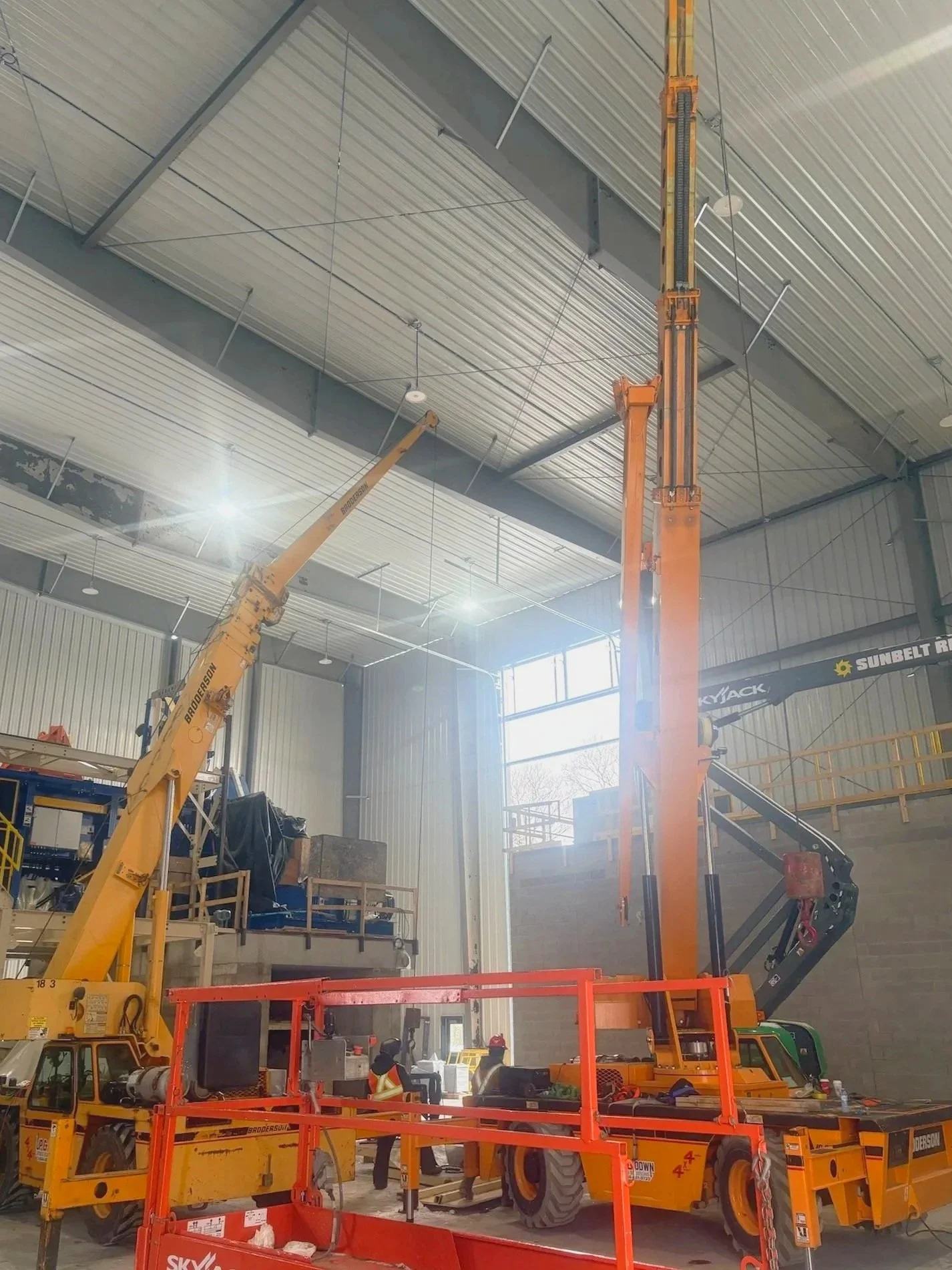 Indoor construction site with two large orange cranes lifting objects, workers in safety vests and helmets, and construction equipment.
