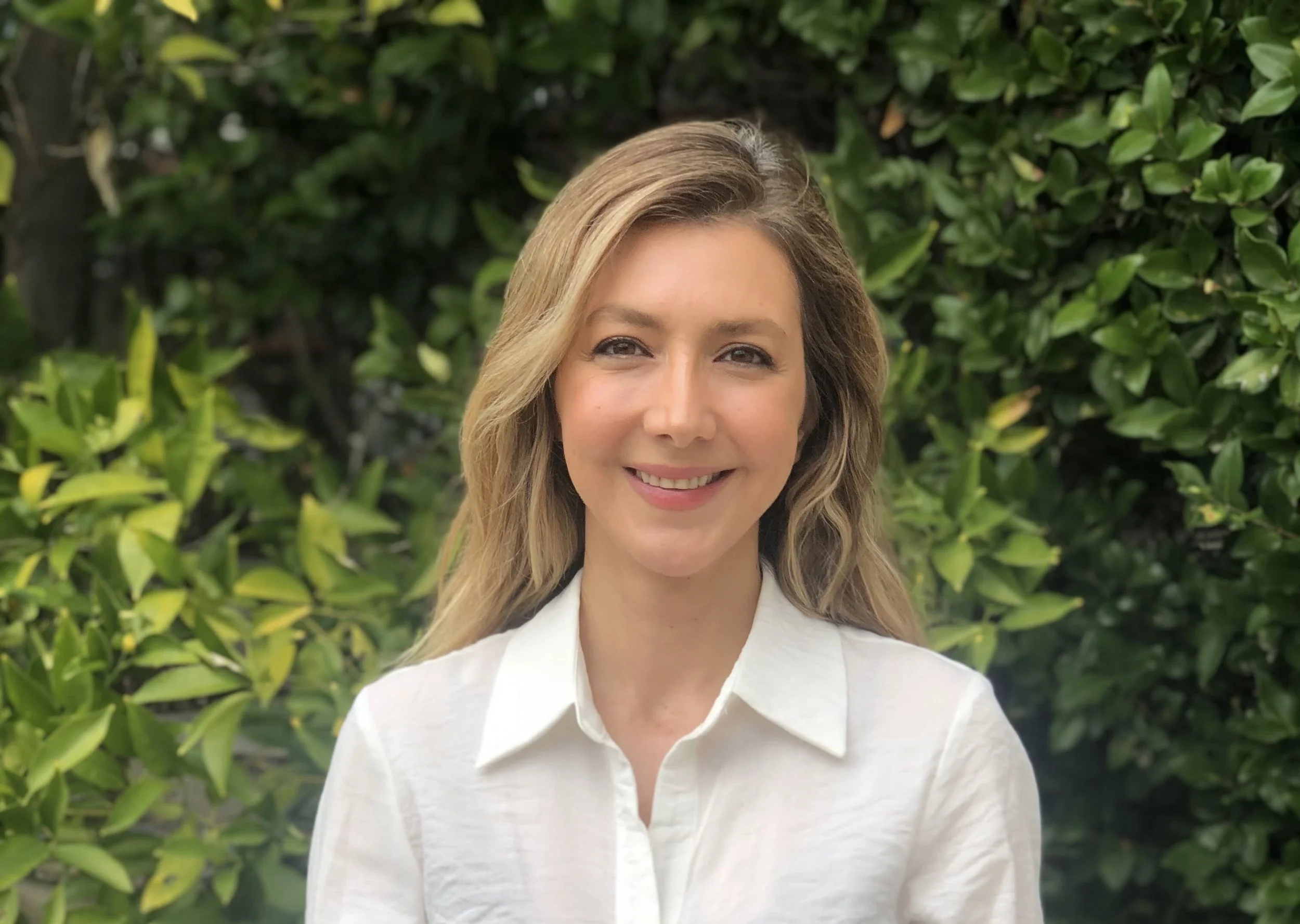 A woman with long, wavy blonde hair smiling and wearing a white button-up shirt, standing outdoors in front of green foliage.