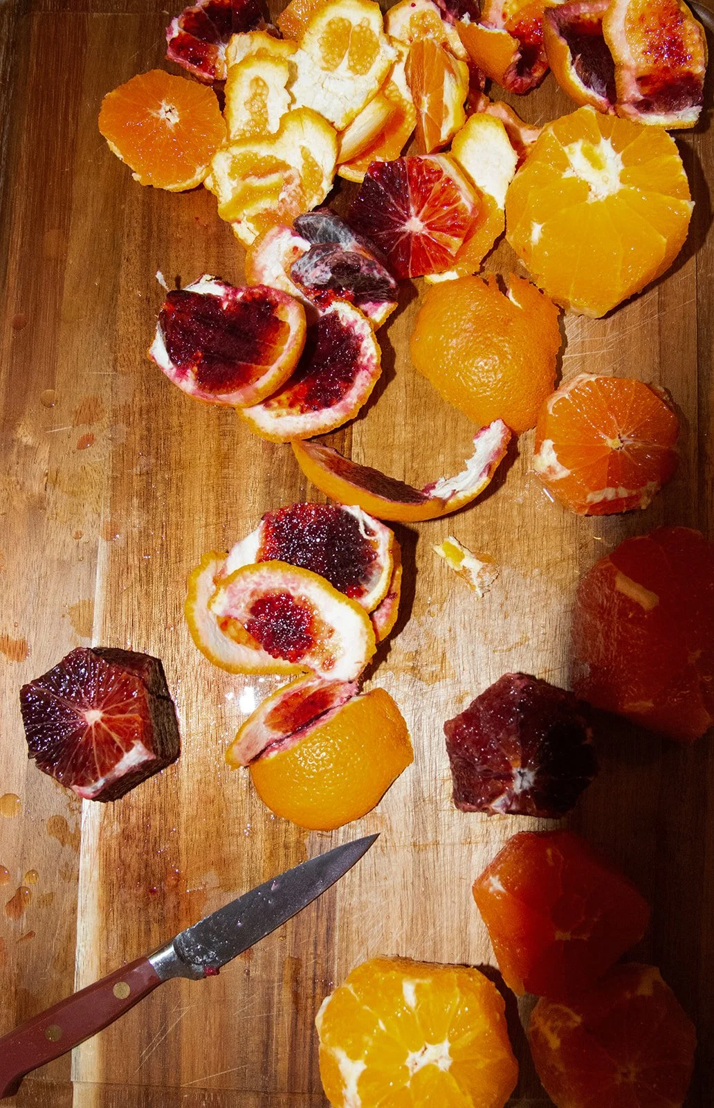 Assorted cut citrus fruits on a wooden cutting board, including oranges, tangerines, and blood oranges, some partially peeled and sliced.