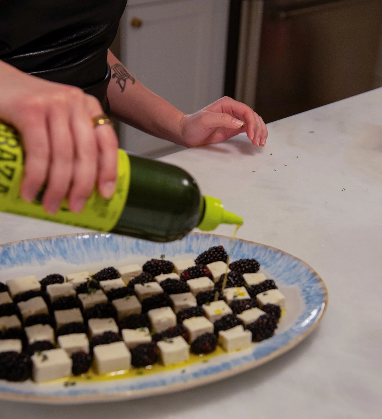 Person drizzling olive oil onto a plate of small cheese cubes and blackberries on a white table.