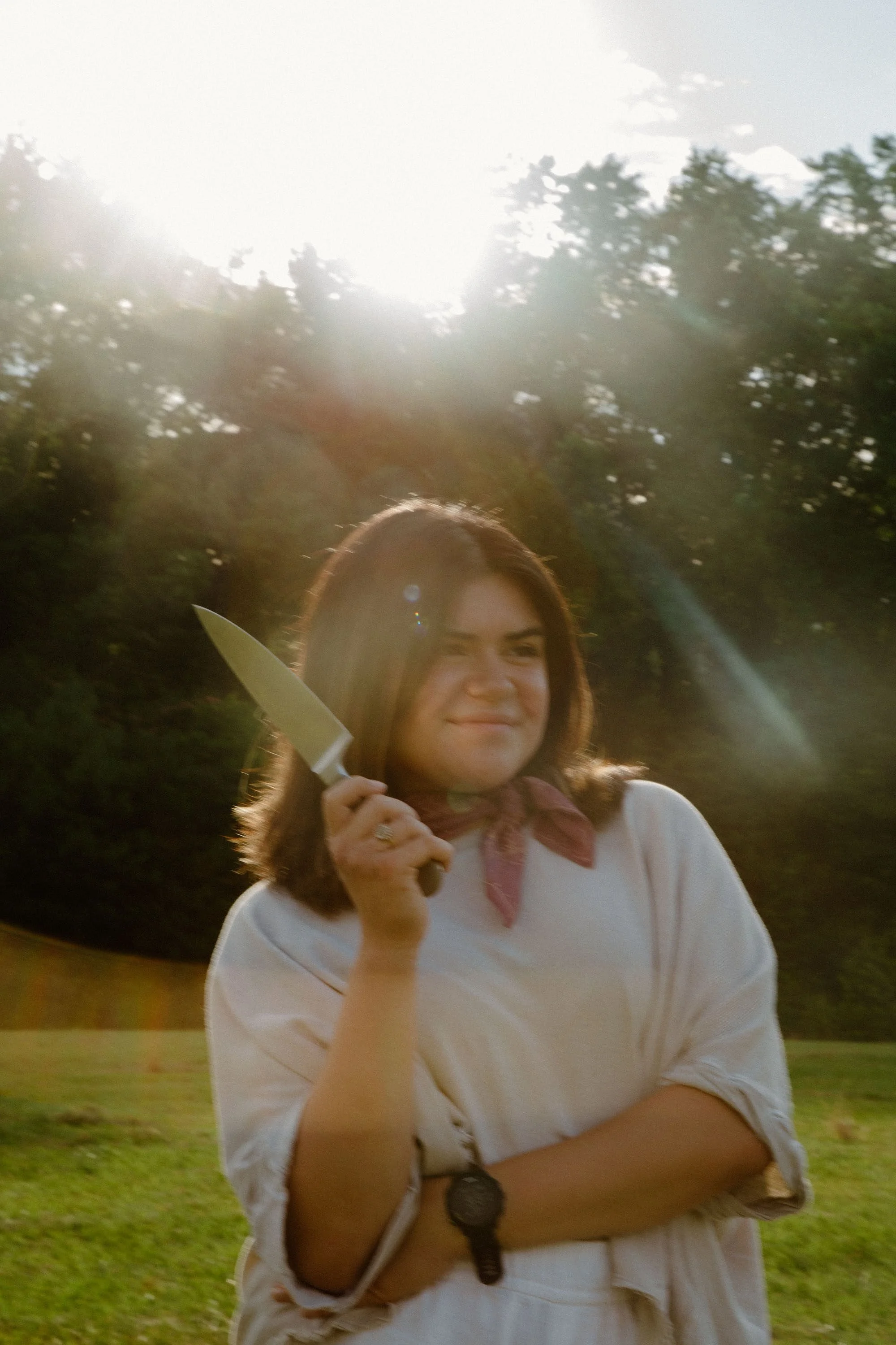 A woman with shoulder-length brown hair, wearing a white shirt and a pink bandana, holds a large kitchen knife in a playful, menacing pose outdoors during daylight, with trees in the background and sunlight creating a lens flare.