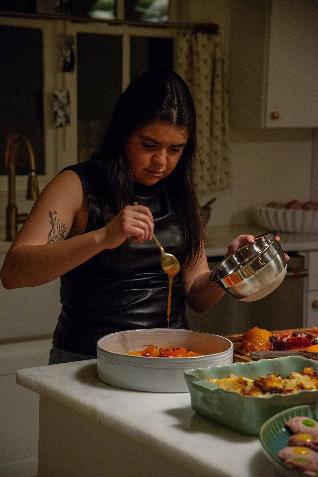 A woman with long dark hair and a tattoo on her upper arm is pouring sauce from a spoon into a dish on a kitchen counter.