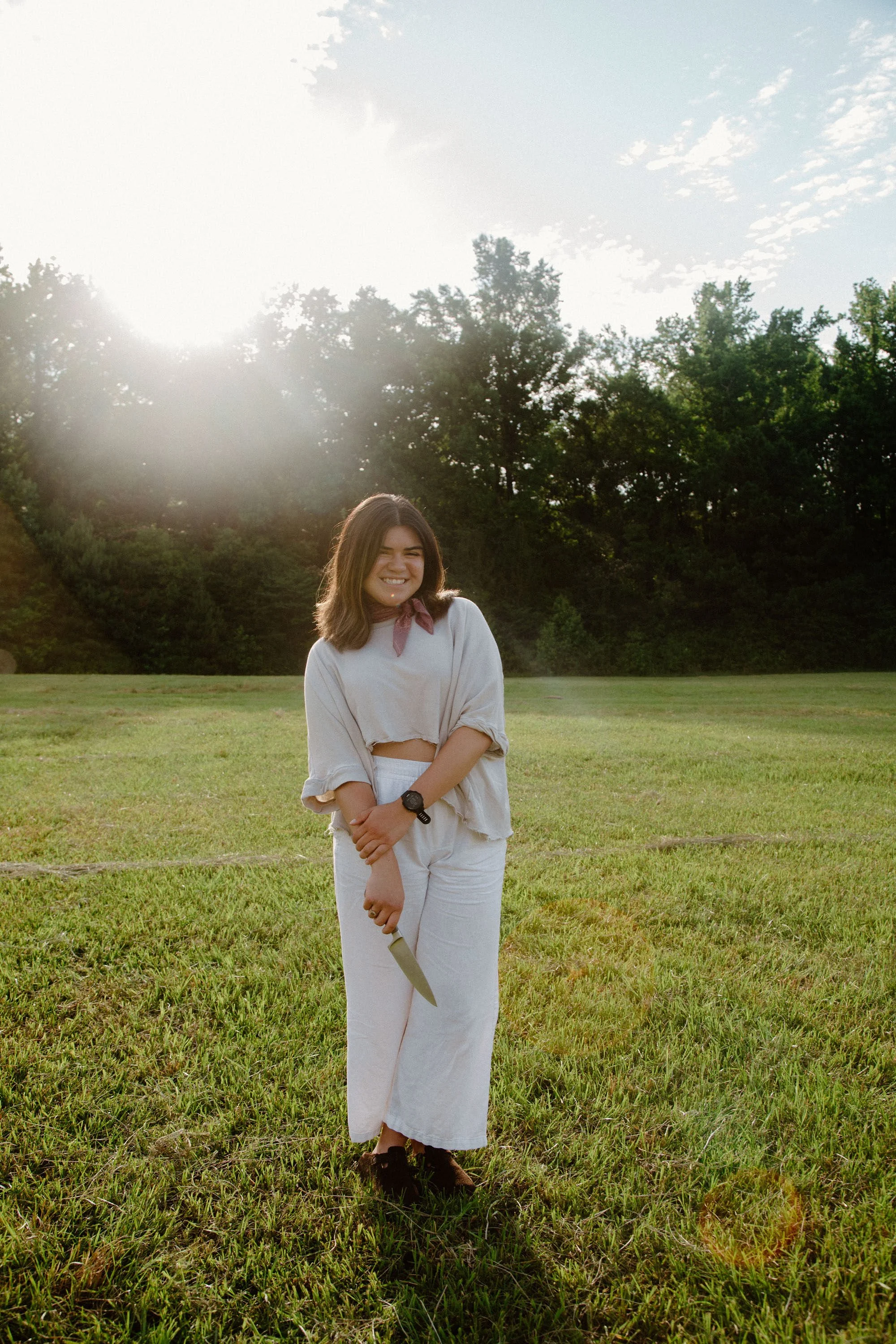 A young woman with shoulder-length brown hair standing in a grassy field, smiling at the camera, with the sun setting behind trees in the background.