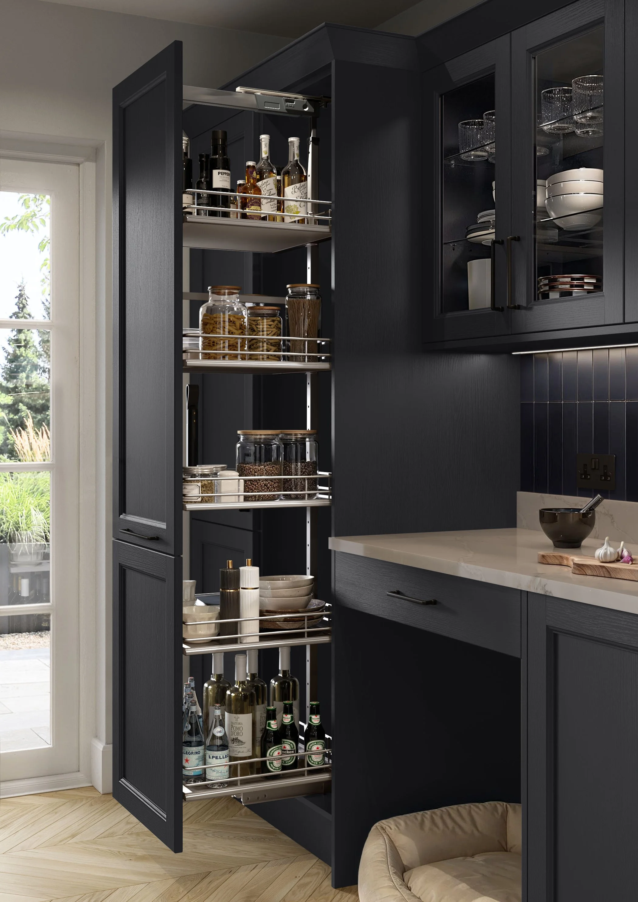 A dark navy kitchen cabinet with a pull-out pantry containing bottles of alcohol, jars of pasta, and condiments. To the right, there are glassware, bowls, and plates stored in the upper cabinet with glass doors. A beige countertop with a small bowl, garlic, and a wooden cutting board is visible in the foreground. To the left, there's a white door with glass panels leading outside to a garden area.