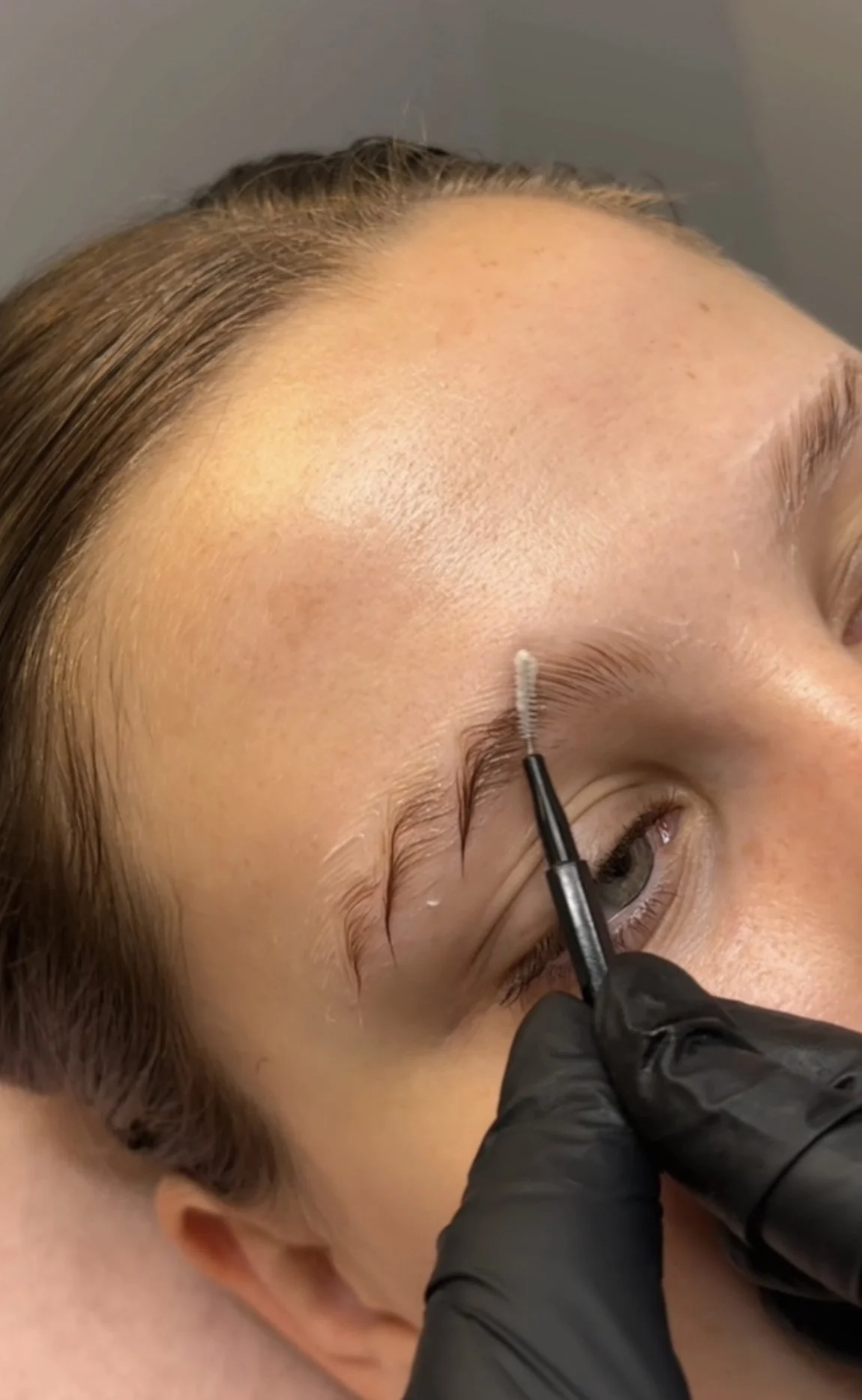Close-up of a person receiving an eyebrow lamination procedure, with a technician wearing black gloves using a small tool to apply pigment to the eyebrow area.