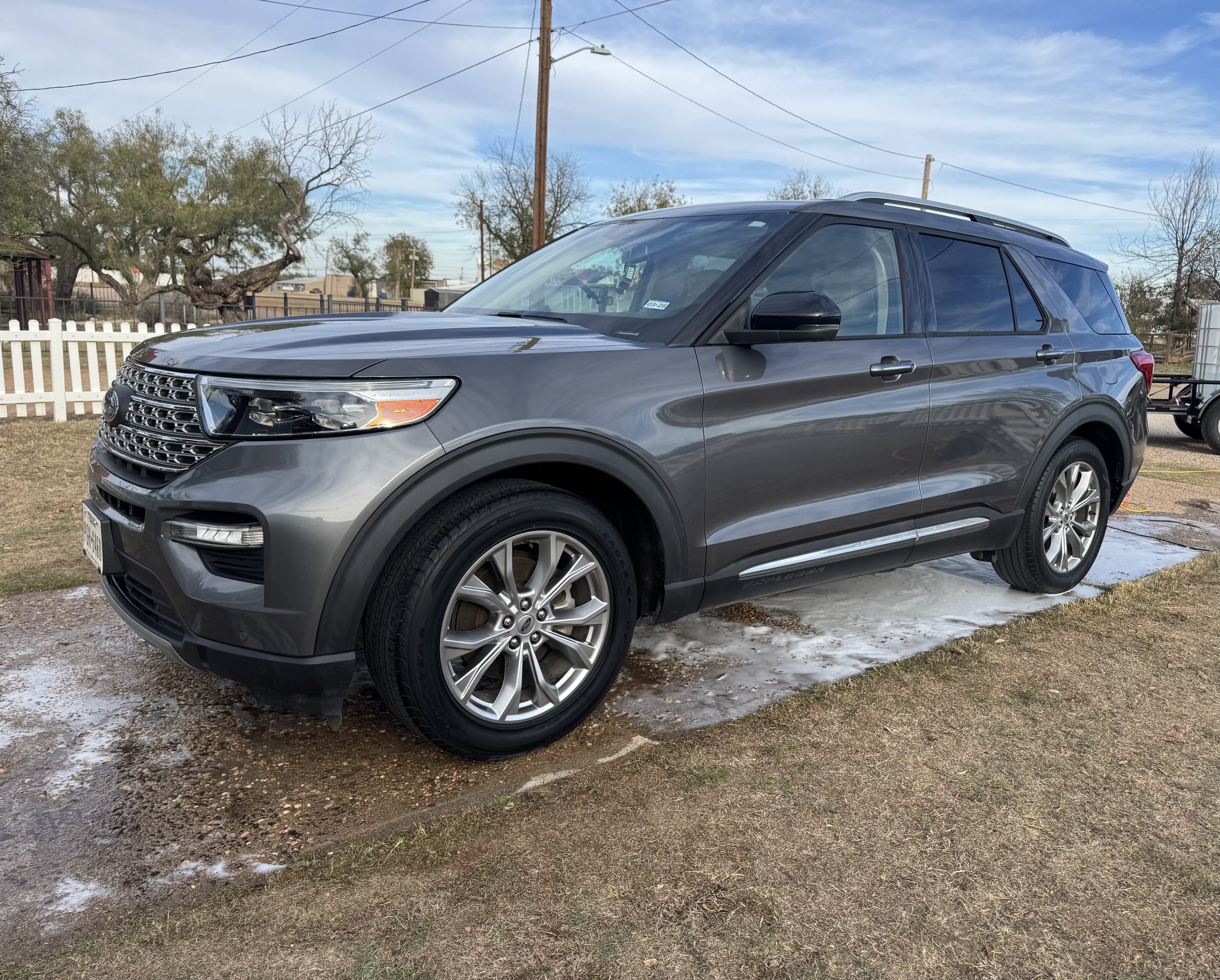 A gray SUV parked on muddy ground with soap suds around the tires in a rural area with a white picket fence and trees in the background under a partly cloudy sky.