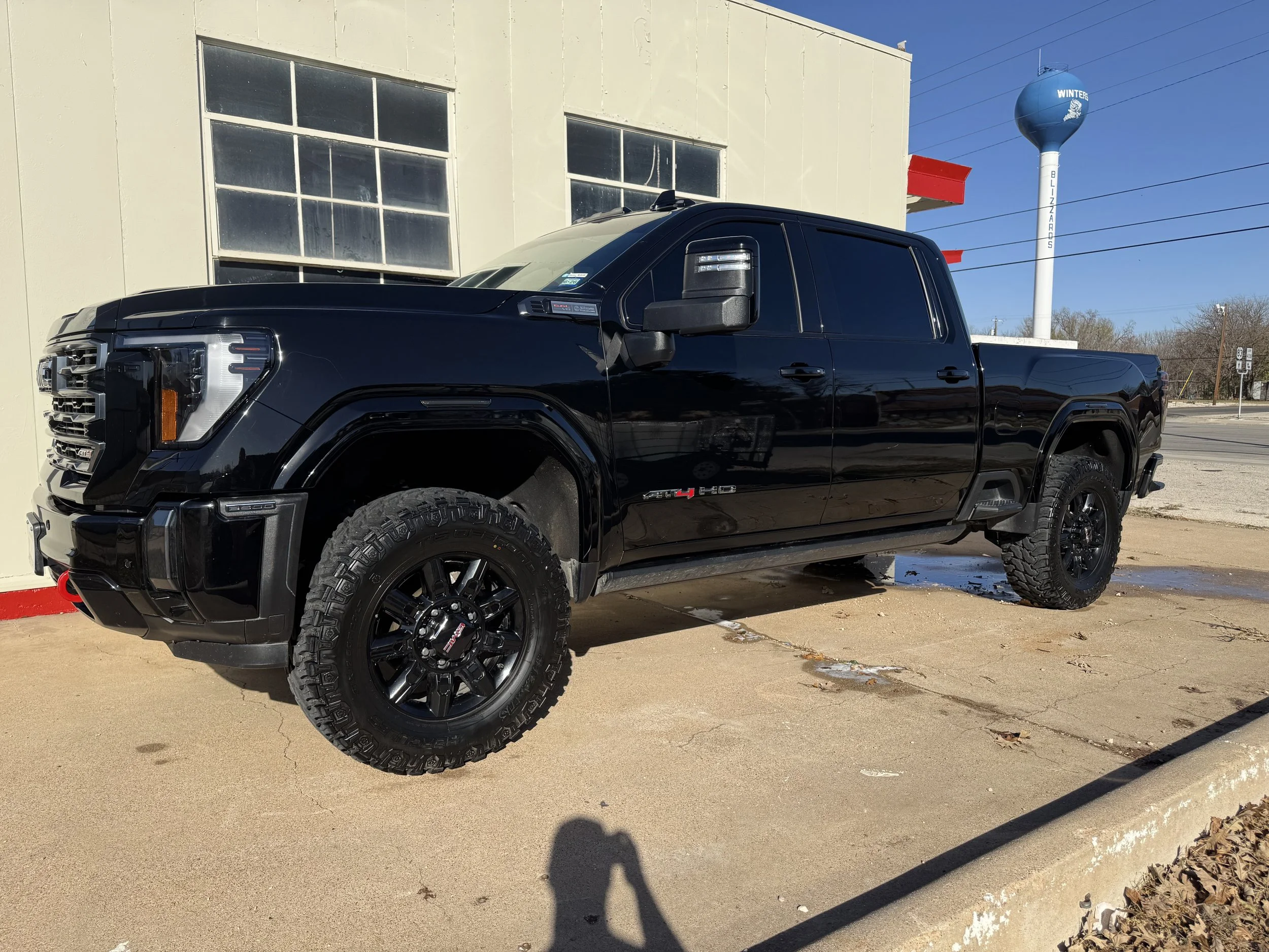 Black GMC pickup truck parked outside a building with large windows, beside a water tower marked "WINTERS" with a snowflake logo, on a sunny day.