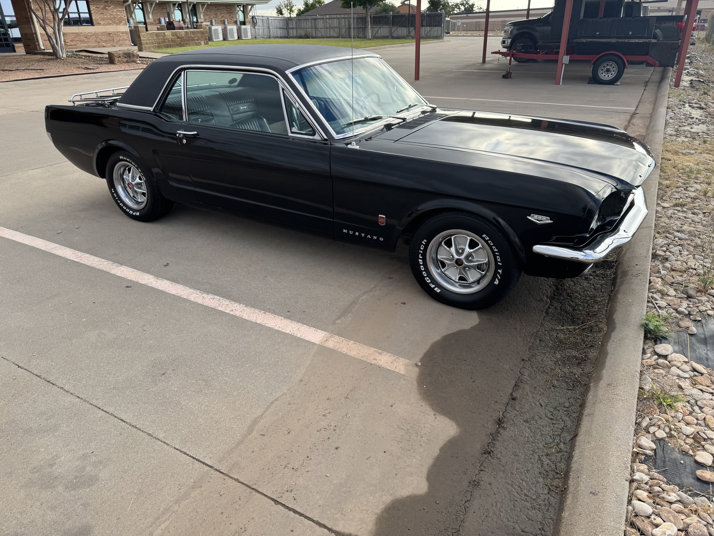 A vintage black Ford Mustang parked in a parking lot next to a curb with rocks on the side. The car has a hardtop with chrome accents and white-letter tires.