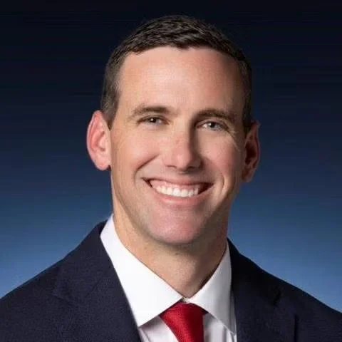 Portrait of a smiling man wearing a suit and red tie, with short dark hair and a dark background.