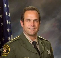A man in a sheriff's uniform standing in front of an American flag.