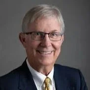 A smiling man in a navy blue suit, white shirt, and red polka dot tie sitting in front of a bookshelf filled with red and blue books, with an American flag standing beside him.