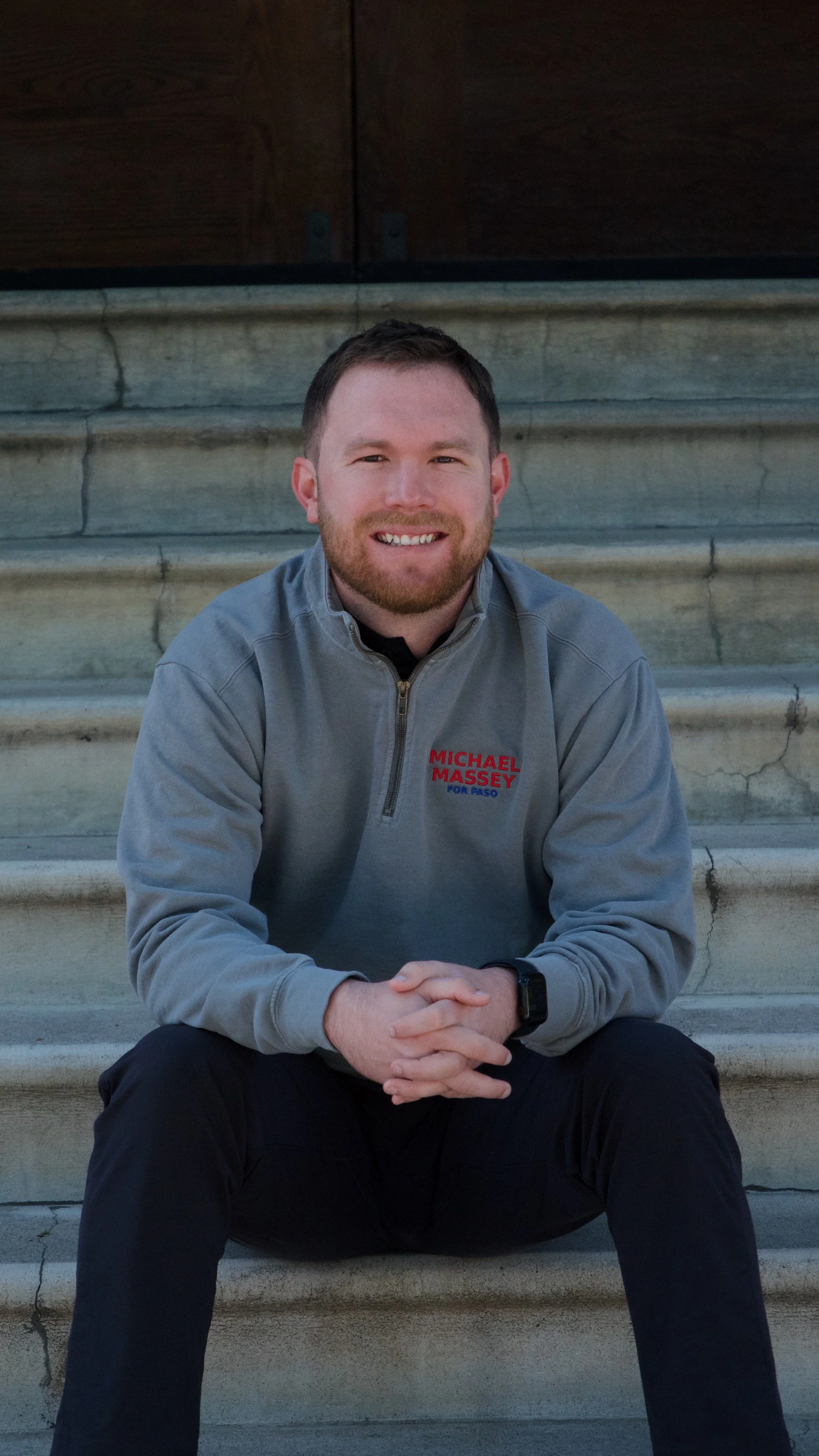 A man sitting on stone steps, wearing a gray pullover with red embroidered text that reads "Michael Massey for Paso." He has short brown hair, a beard, and is smiling at the camera.