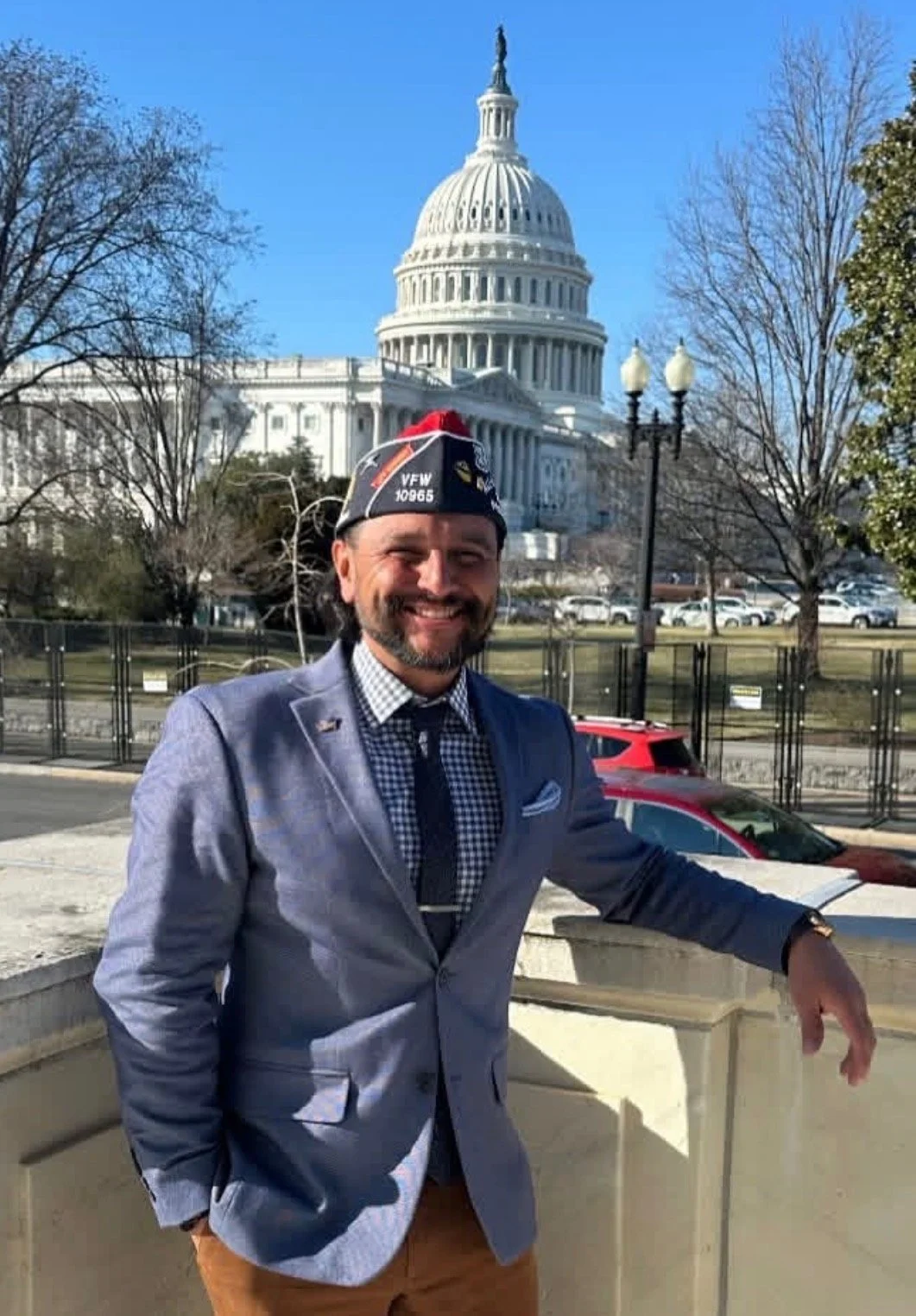 A man smiling in front of the United States Capitol building, wearing a dark suit, a checkered shirt, a necktie, and a military-style hat with insignia.