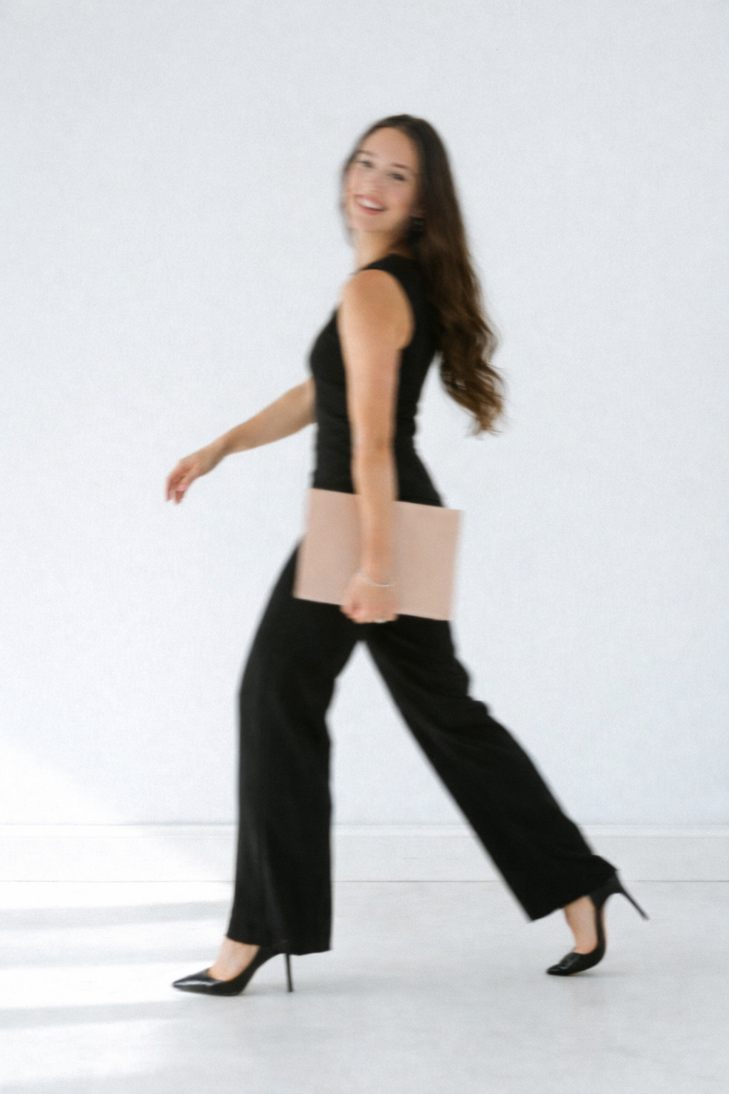 Woman walking, smiling, holding a beige clutch, wearing black sleeveless top, black pants, and black high heels, with long wavy brown hair, against a plain light background.