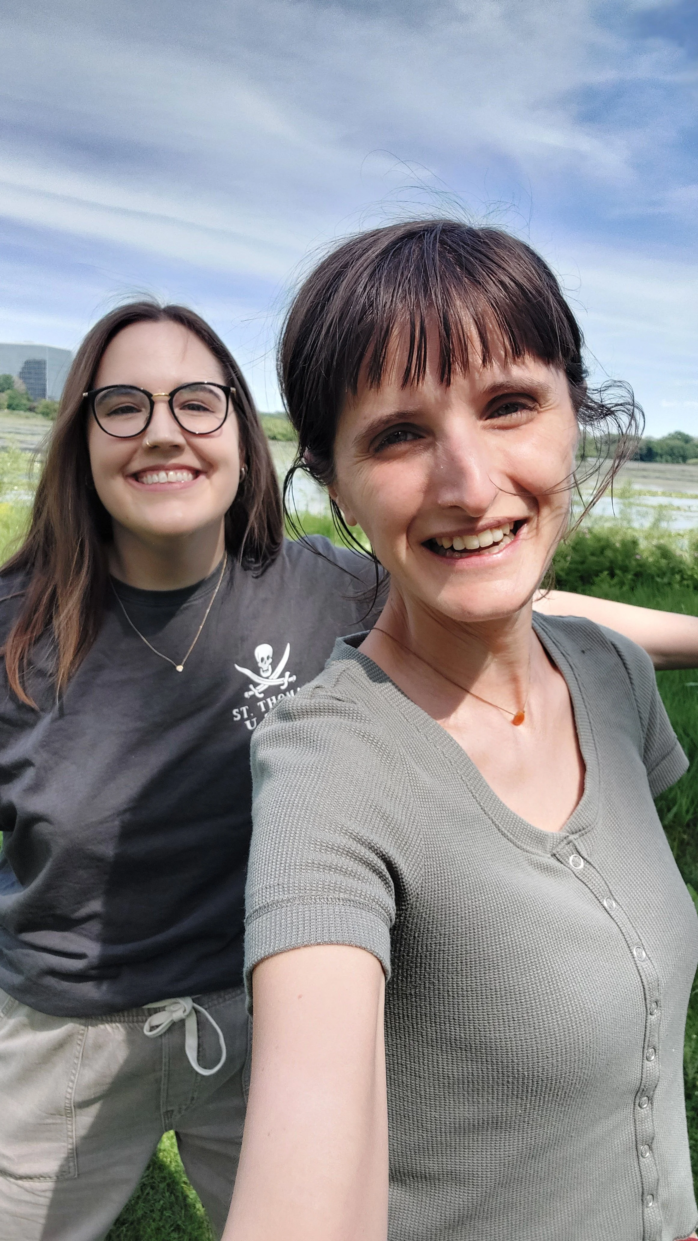 Two women smiling outdoors on a cloudy day, standing close together with greenery and water in the background.