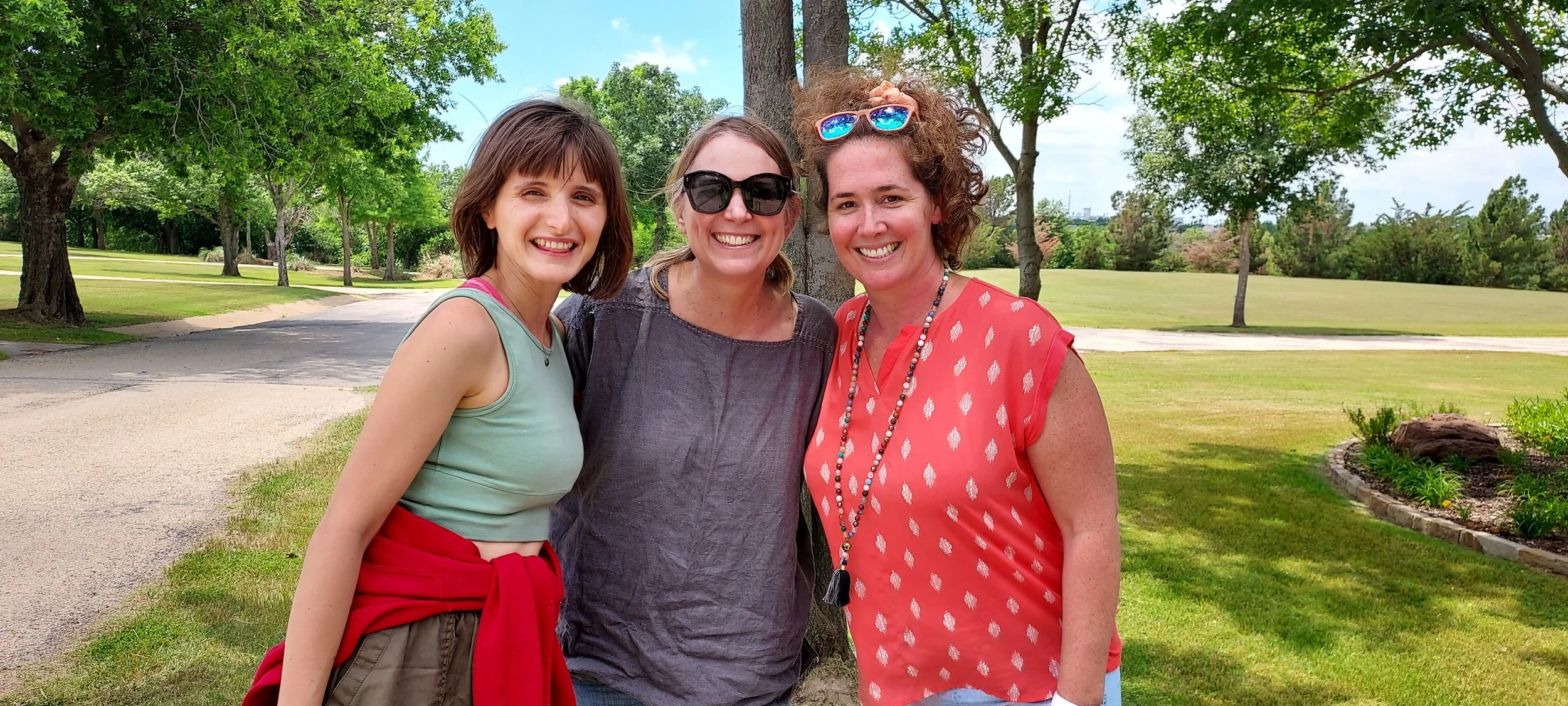 Three women smiling outdoors in a park with trees and grass.