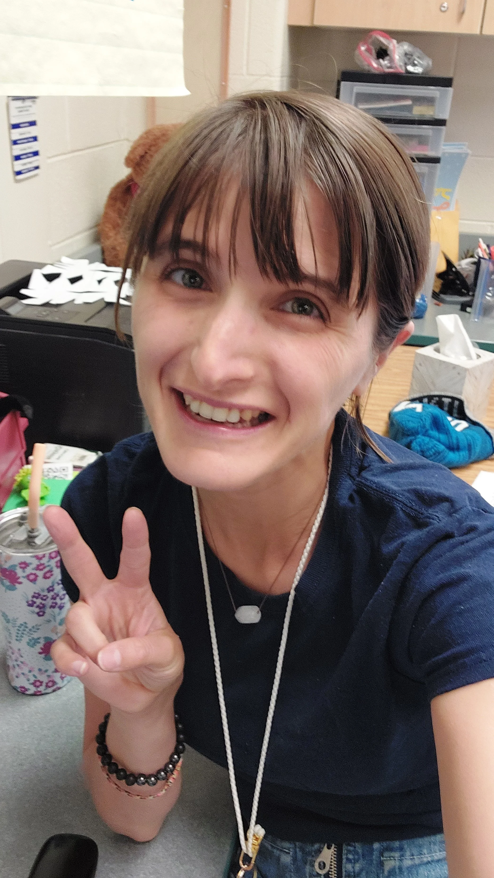 A woman with short brown hair and blue eyes smiling and holding up a peace sign. She is in a classroom or office with various supplies and a stuffed animal in the background.