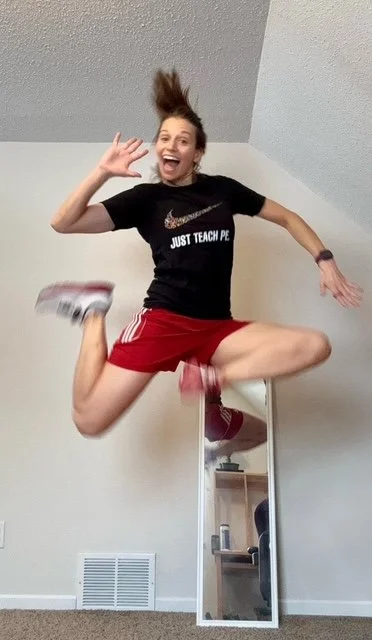 A woman jumping in the air with a big smile, wearing a black Nike t-shirt, red shorts, and sneakers, indoors near a mirror and a vent on the wall.