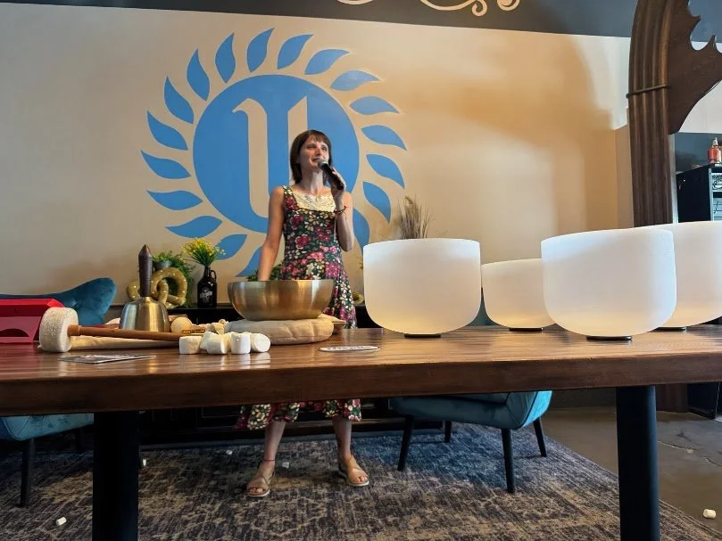 A woman in a floral dress is speaking into a microphone behind a table with singing bowls and a mallet, in front of a wall with a blue logo and large white bowls.