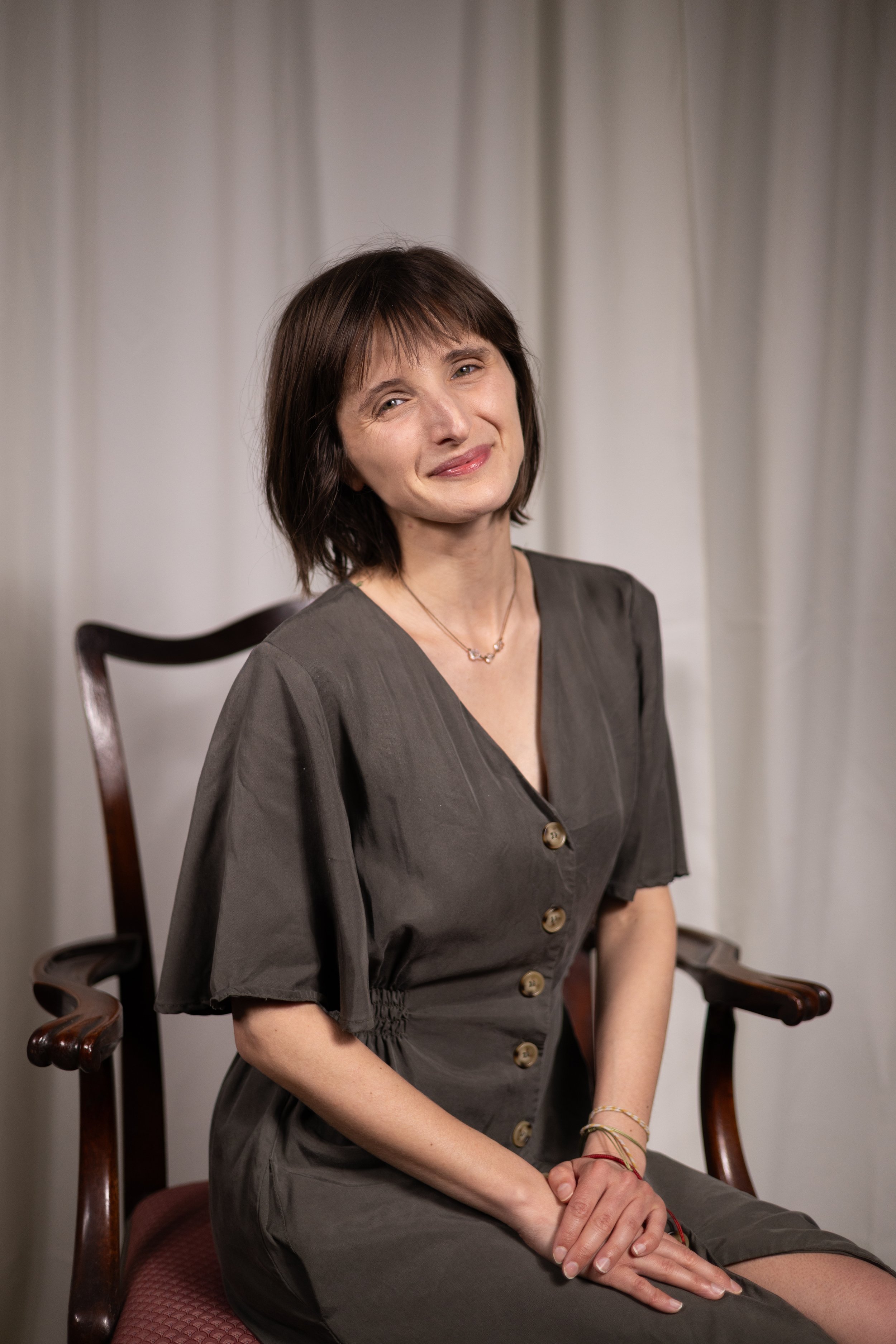 A woman with short brown hair and a slight smile, sitting on a wooden armchair with armrests, against a background of white curtains, wearing a dark gray dress with buttons and jewelry.