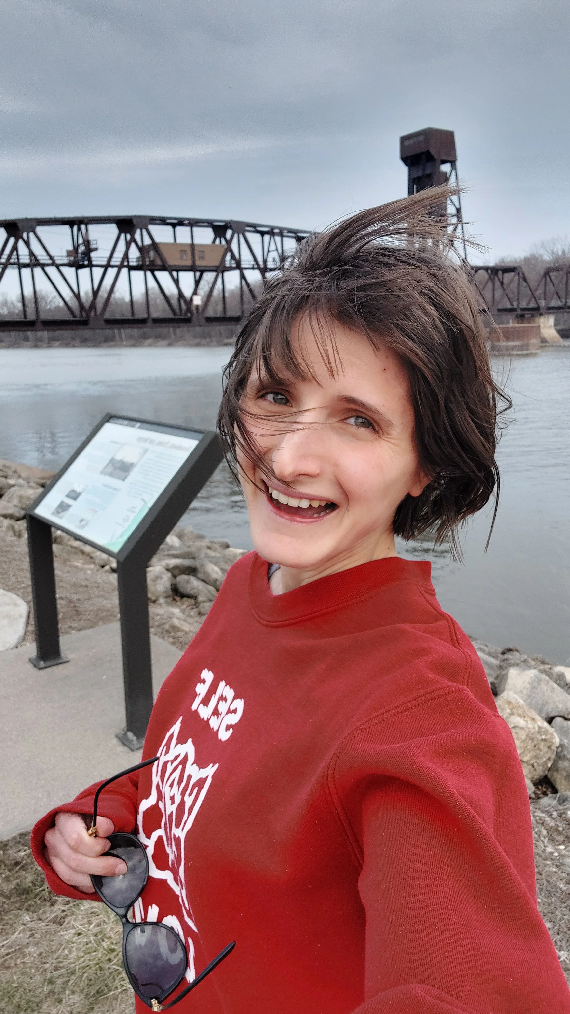 A woman with short, tousled brown hair smiling outdoors near a river, holding sunglasses, with a historic train bridge and an informational sign in the background.