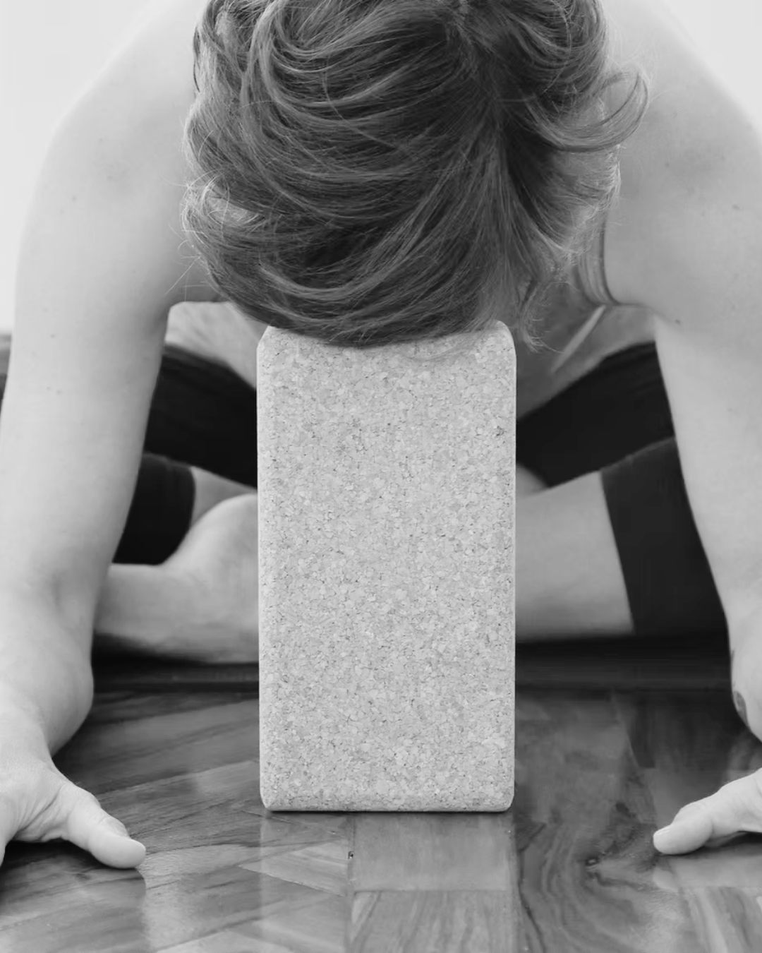 A person practicing yoga in a child’s pose position with their forehead resting on a yoga block on a wooden floor.
