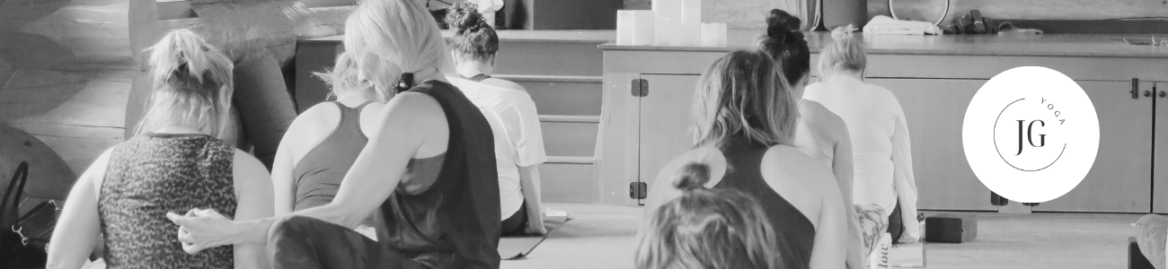 Group of women in yoga class sitting on mats, facing away from the camera, with a logo that says "JG Yoga" in a circle in the upper right corner.