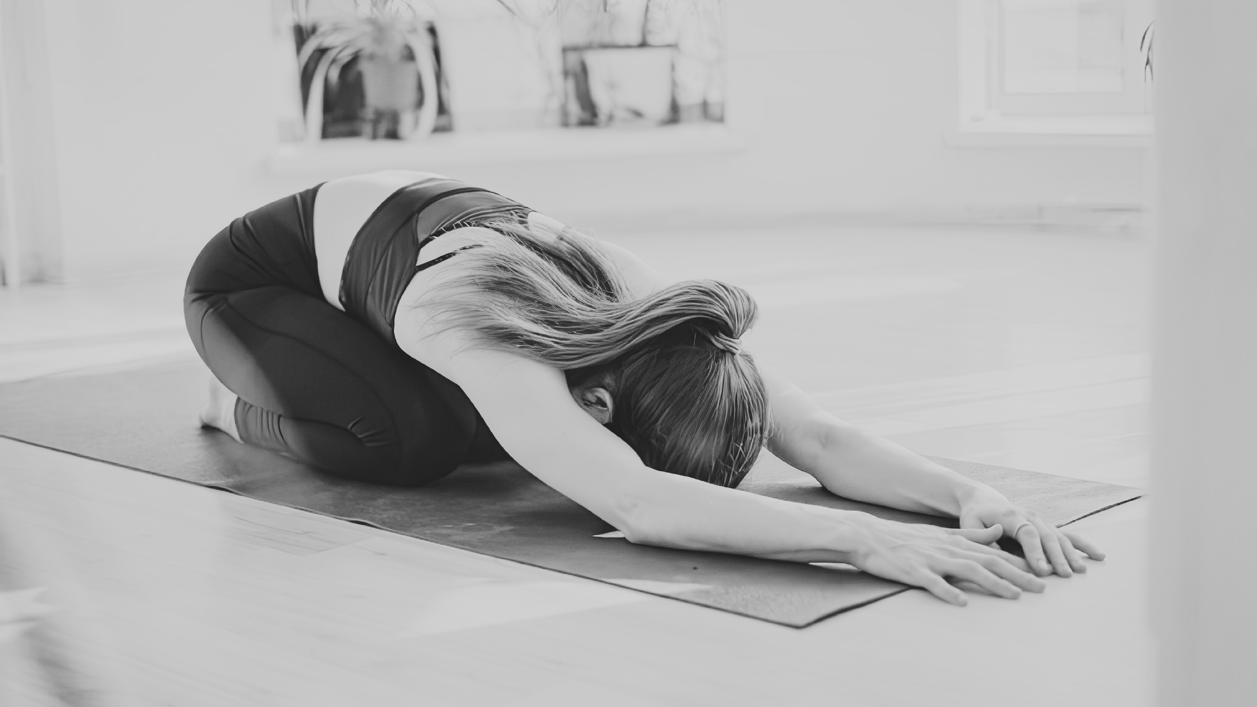 A person practicing yoga in a child's pose position on a yoga mat in a bright room with potted plants on the window sill.