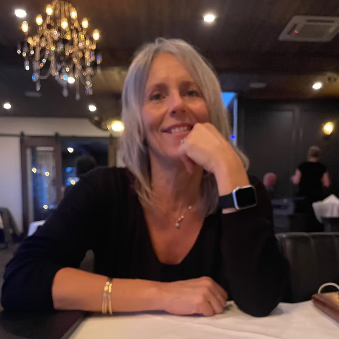 Jen Dudar with gray hair sitting at a table in a dimly lit restaurant, smiling, with a chandelier above her and other diners in the background.