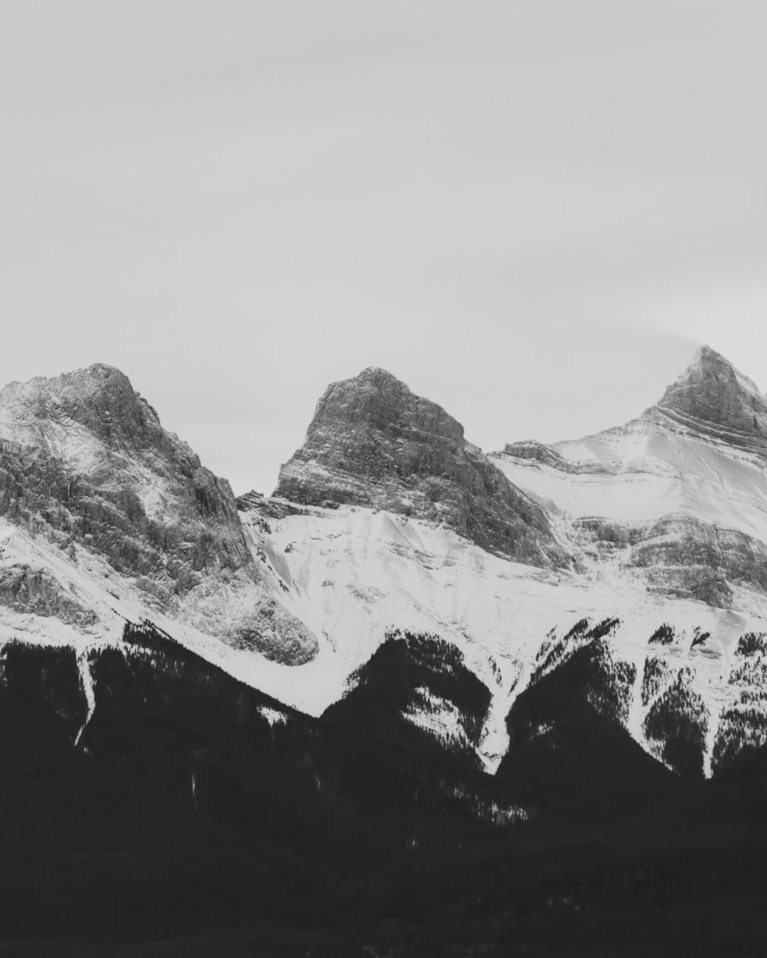 Black and white photograph of snow-covered Three Sisters mountain range in Canmore, Alberta, showing their peaks with a cloudy sky in the background.