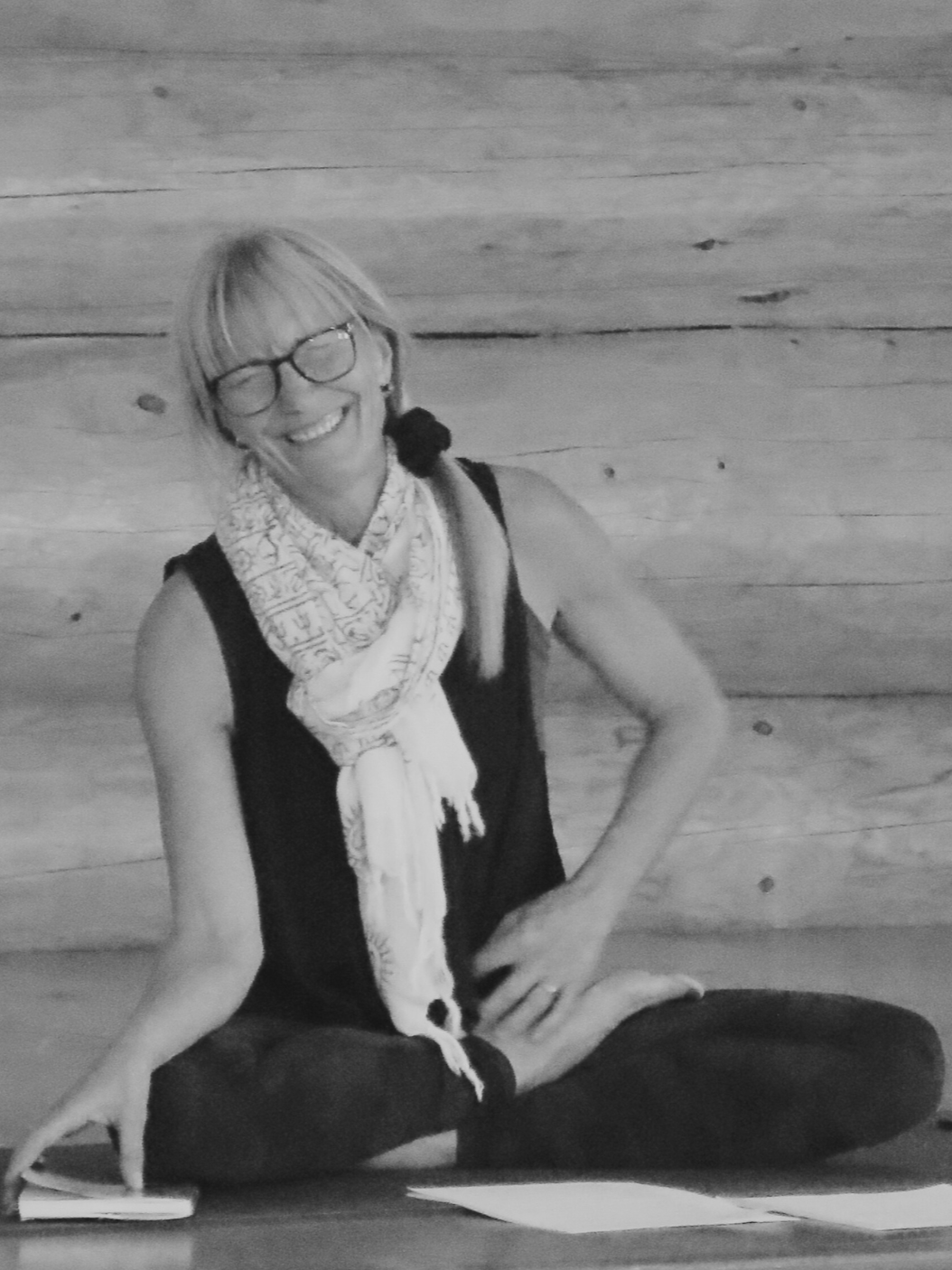 Smiling woman with glasses, wearing a scarf and black sleeveless top, sitting cross-legged on the floor against a wooden wall.