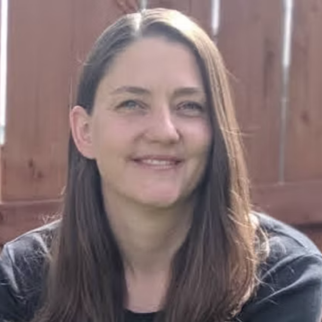 Shannon McKenzie, guest yoga teacher, smiling with long brown hair and blue eyes sitting outdoors in front of a wooden fence.
