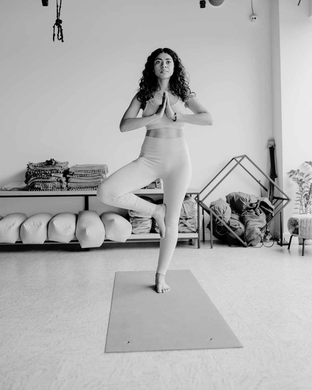 A woman practicing yoga indoors, standing on one leg in a tree pose with hands in prayer position, on a yoga mat in a minimalist room.