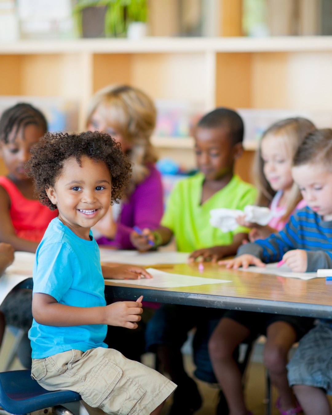 A classroom of young children, with one smiling girl in a blue shirt sitting at a table, surrounded by other kids engaged in activities.