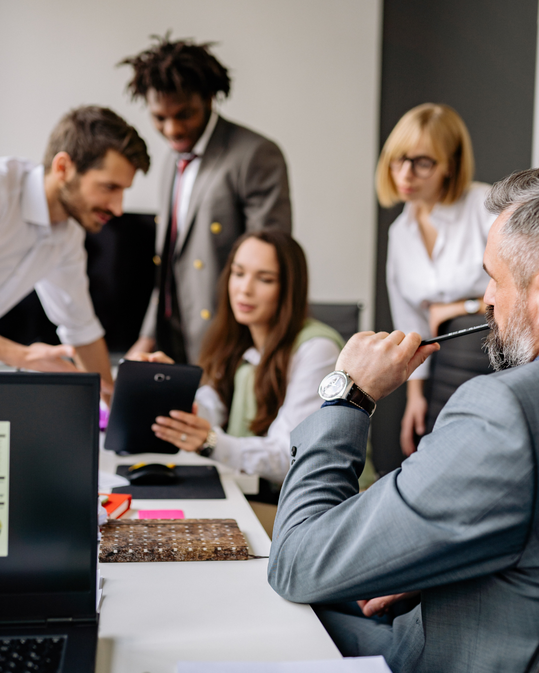 Business team in a meeting discussing around a table, with one woman holding a tablet and a man in a gray suit with a beard listening thoughtfully.