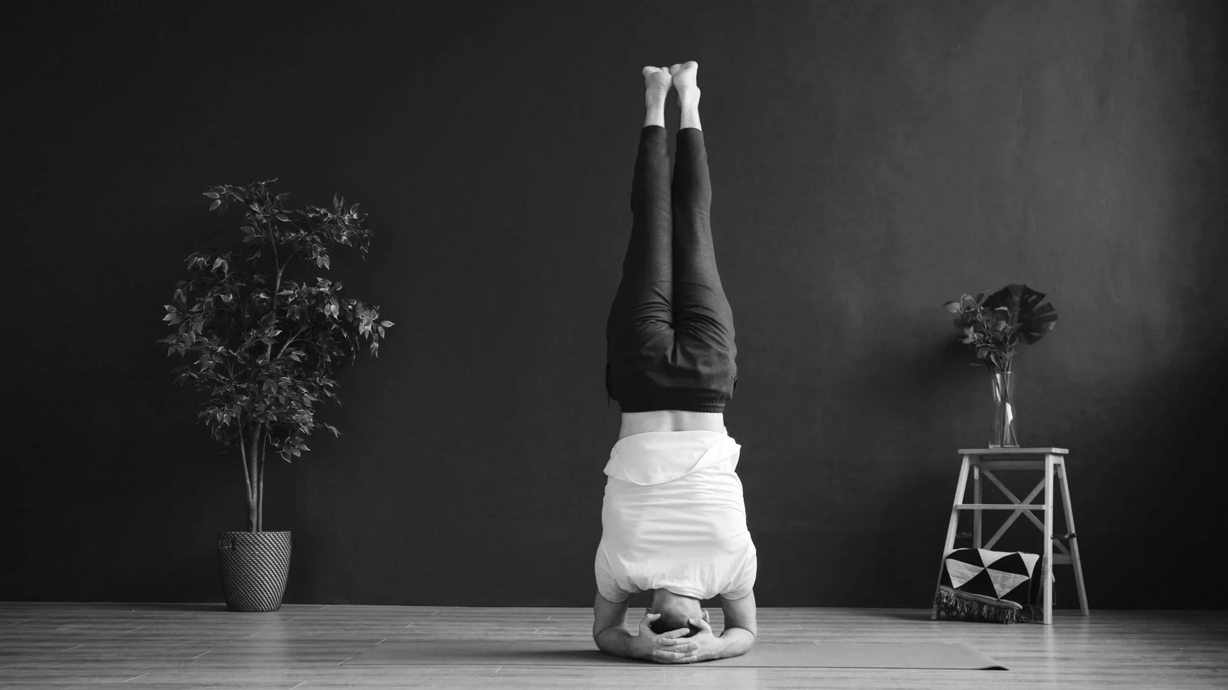 A person doing a headstand yoga pose on a mat in a room with two potted plants and a small table.