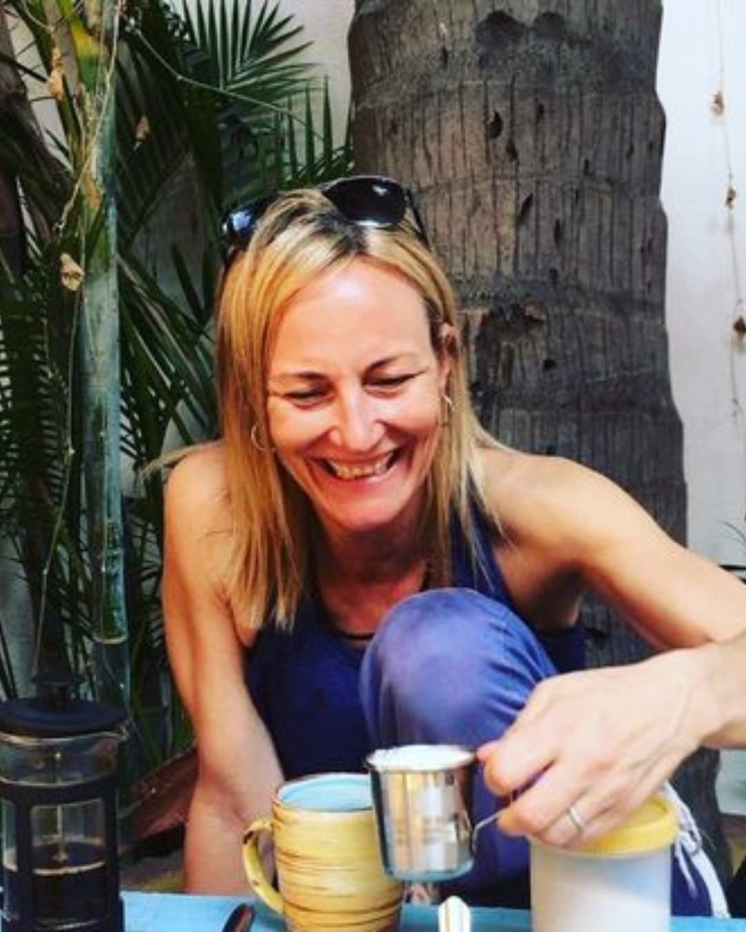 Jan Goranson, Authorized yoga instructor, with blonde hair, wearing sunglasses on her head, smiling while pouring a hot drink from a small metal pot into a yellow mug at an outdoor setting with green plants and a wooden background.