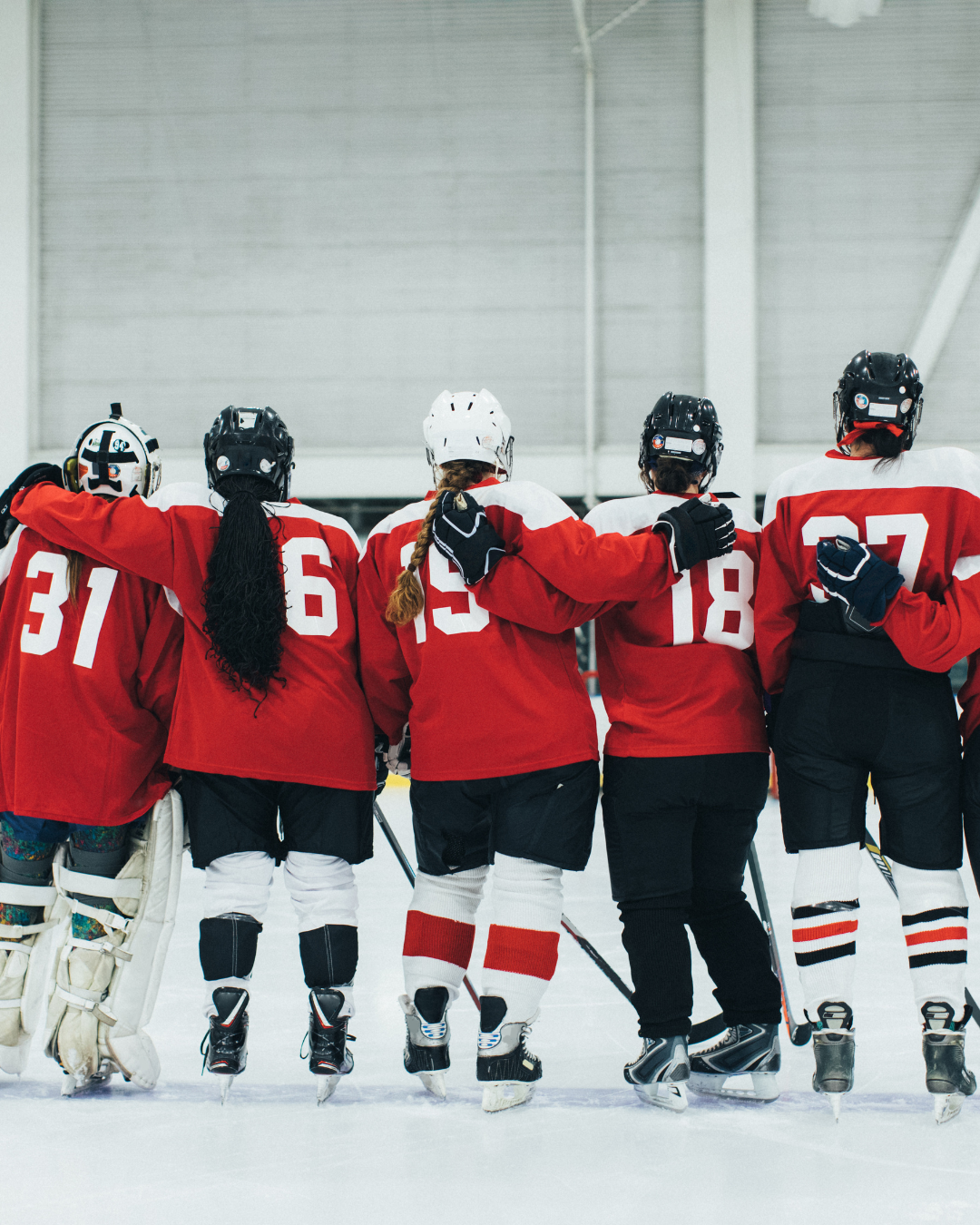 Girls hockey team standing together on ice, with arms around each other's shoulders, wearing red and white jerseys and helmets, in an indoor hockey rink.